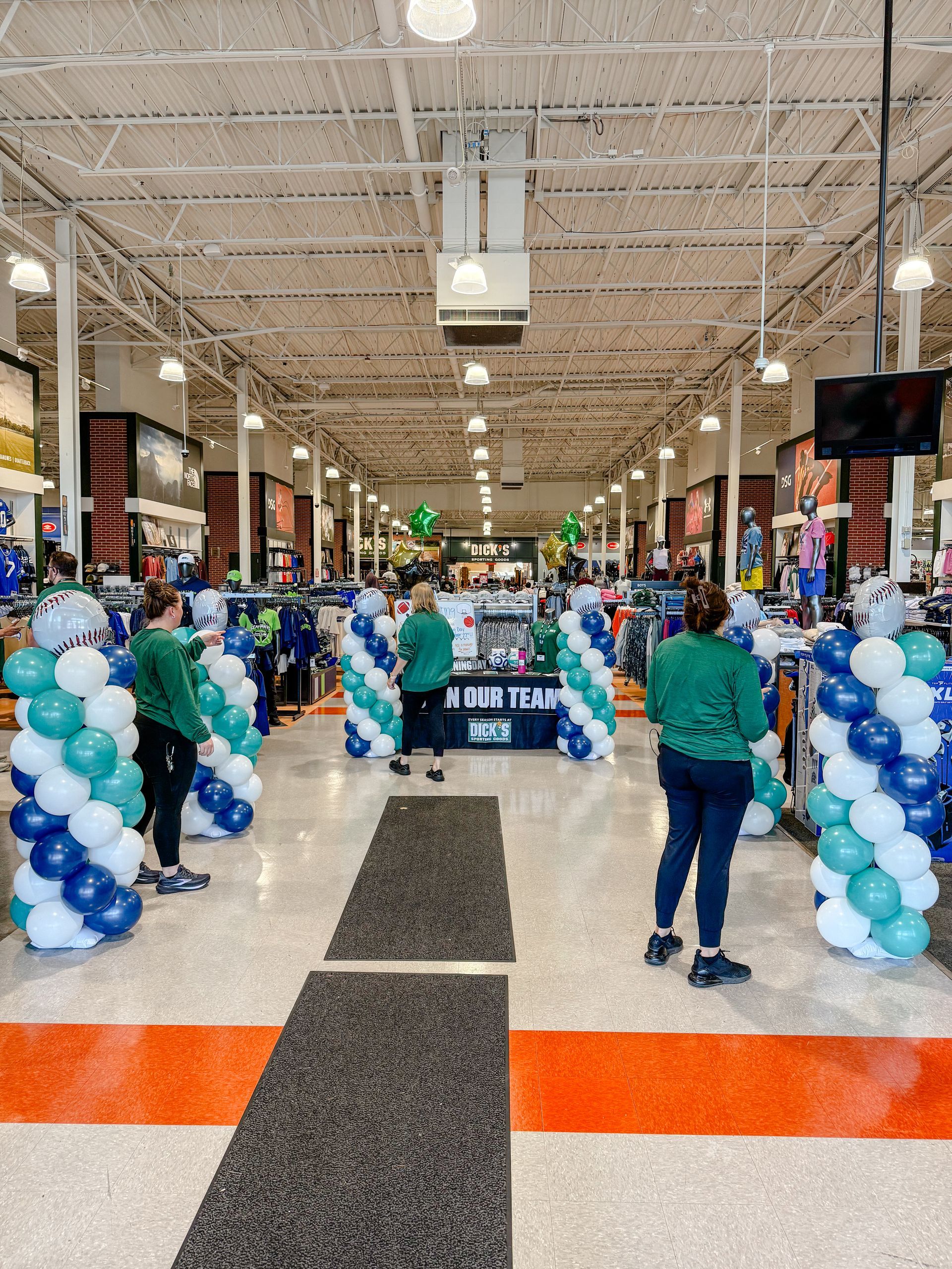 Inside a sporting goods store, people in green shirts stand near balloon columns at a store entrance.