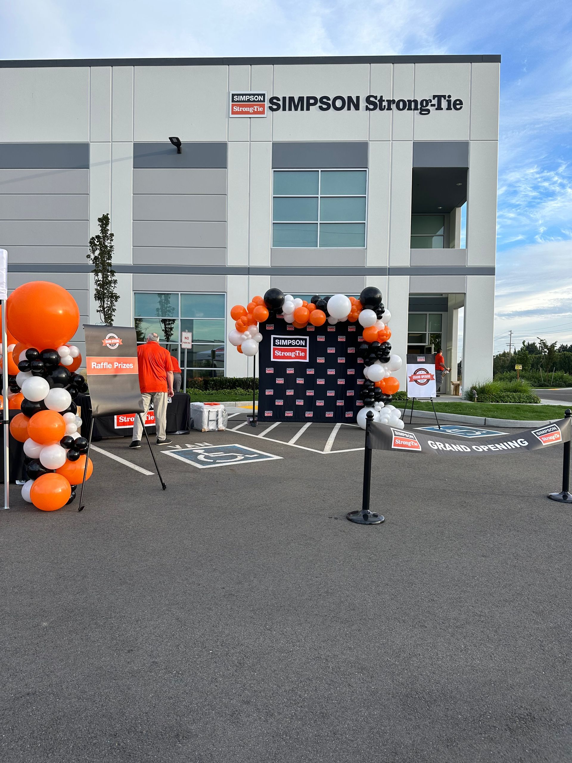 An outdoor event with orange and black balloons, a step-and-repeat backdrop, and a person standing in a parking lot.