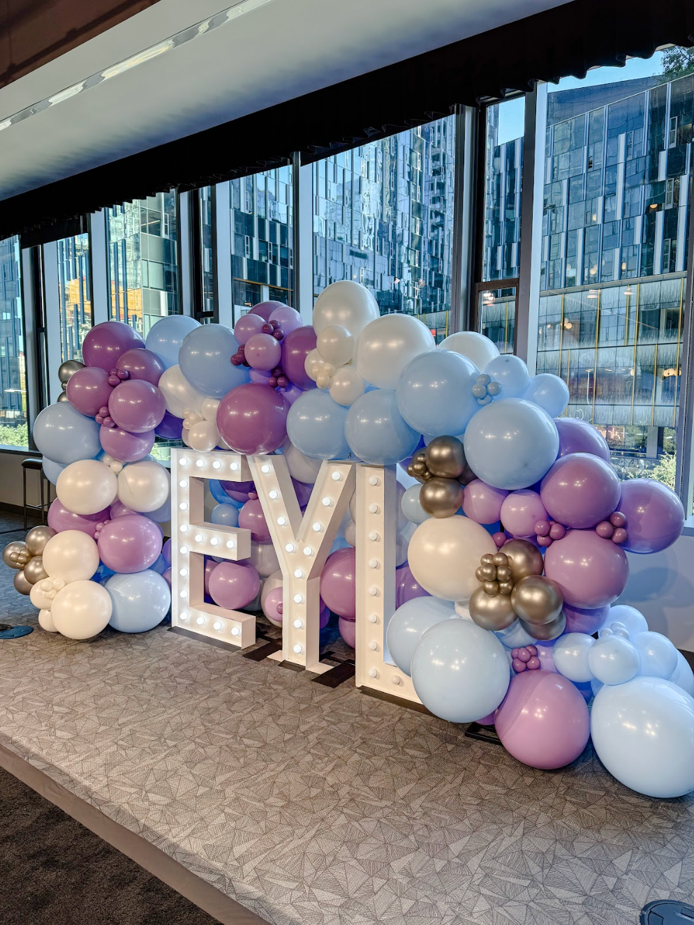 A balloon garland in shades of blue, purple, white, and gold draped over giant, illuminated marquee letters spelling EYL.