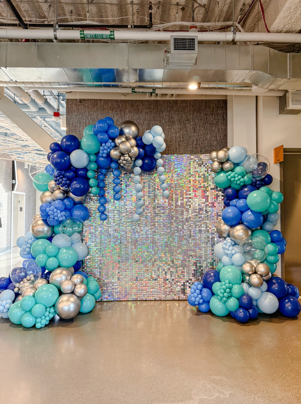 Blue, teal, and silver balloon arch with backdrop of shiny silver squares for a celebration.