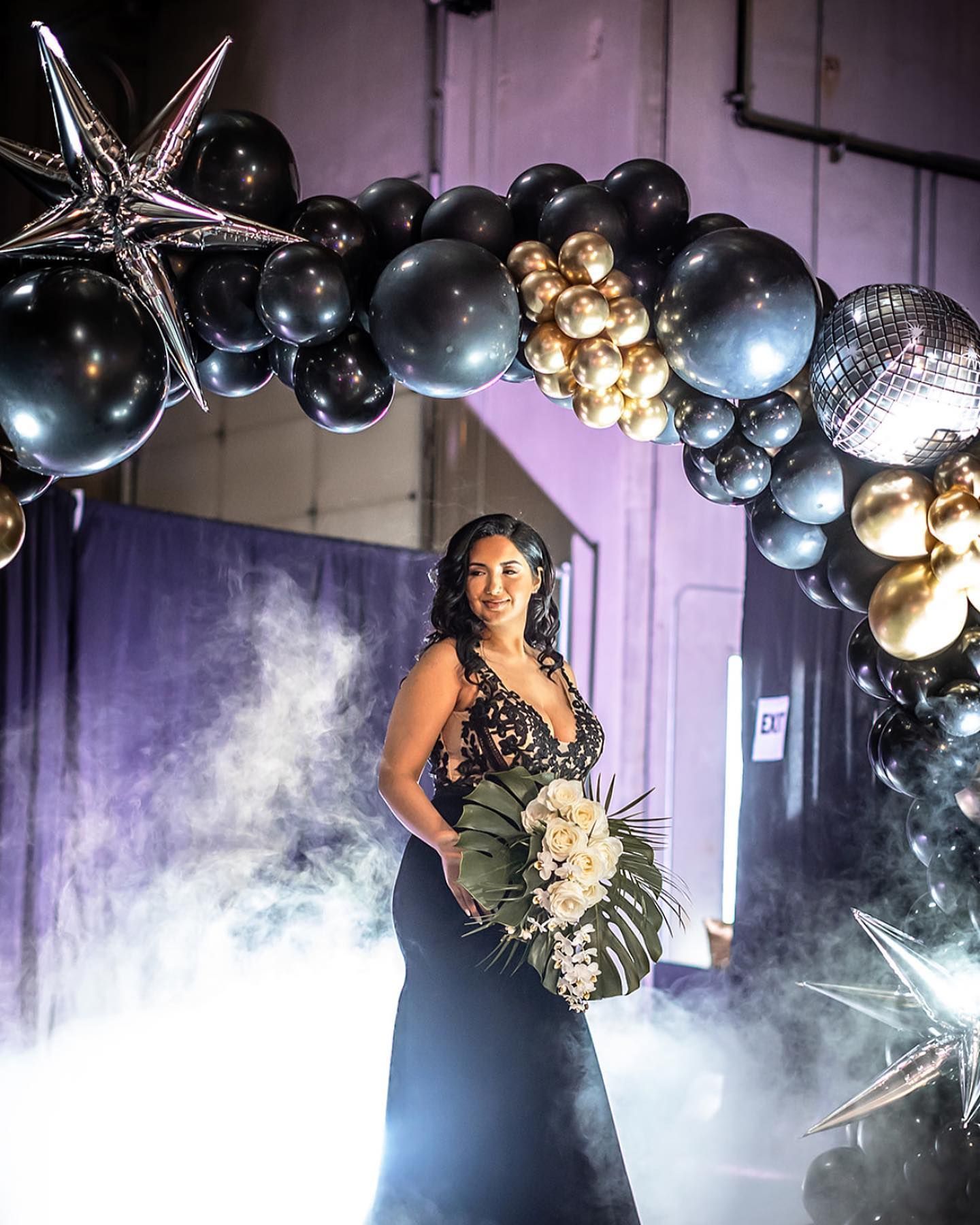 A woman in a black dress is standing in front of a balloon arch on a stage with fog.