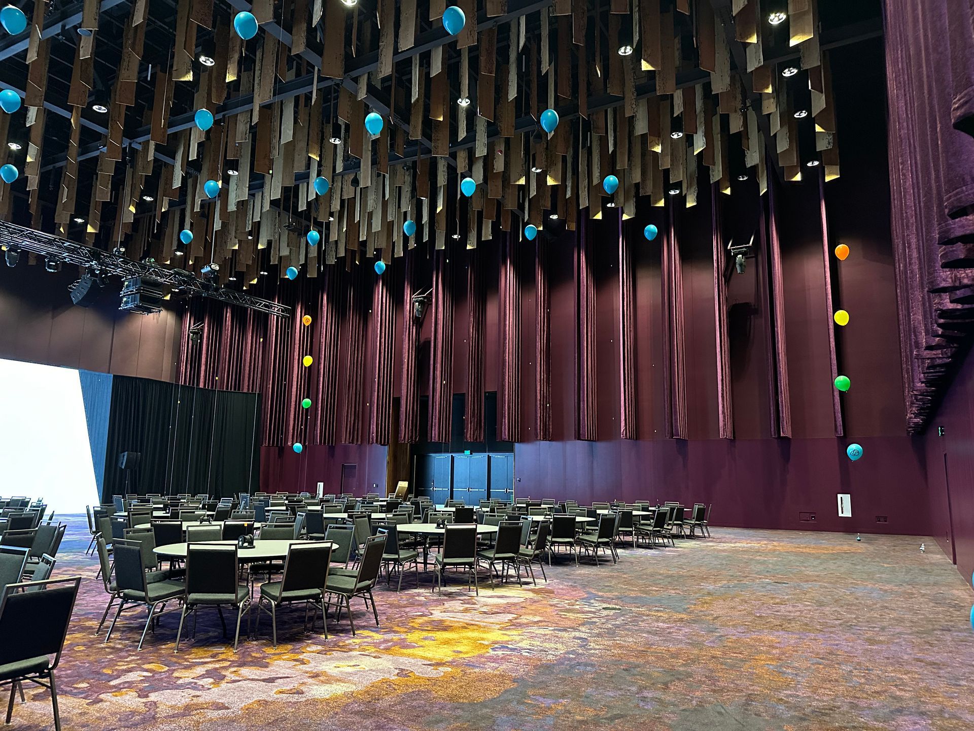 A large auditorium with tables and chairs and balloons hanging from the ceiling.