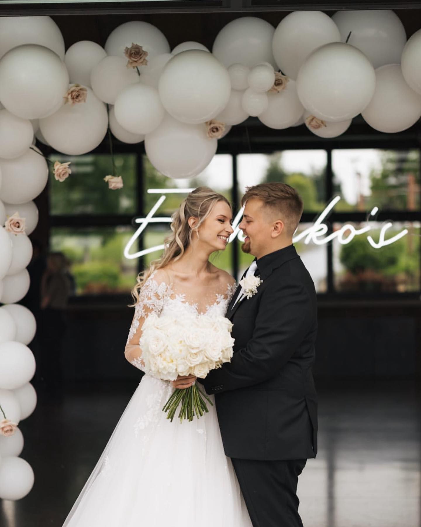 A bride and groom are standing under a white balloon arch.
