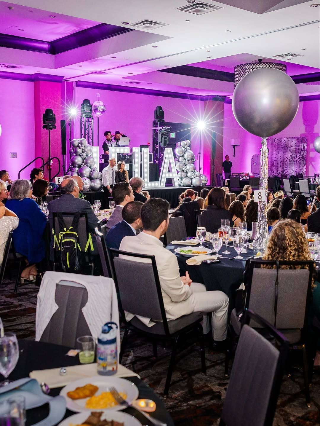 A large auditorium with tables and chairs and balloons hanging from the ceiling.