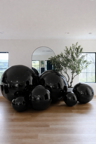 A minimalist room with a light wood floor, an arched mirror, an olive tree, and several glossy black decorative spheres.