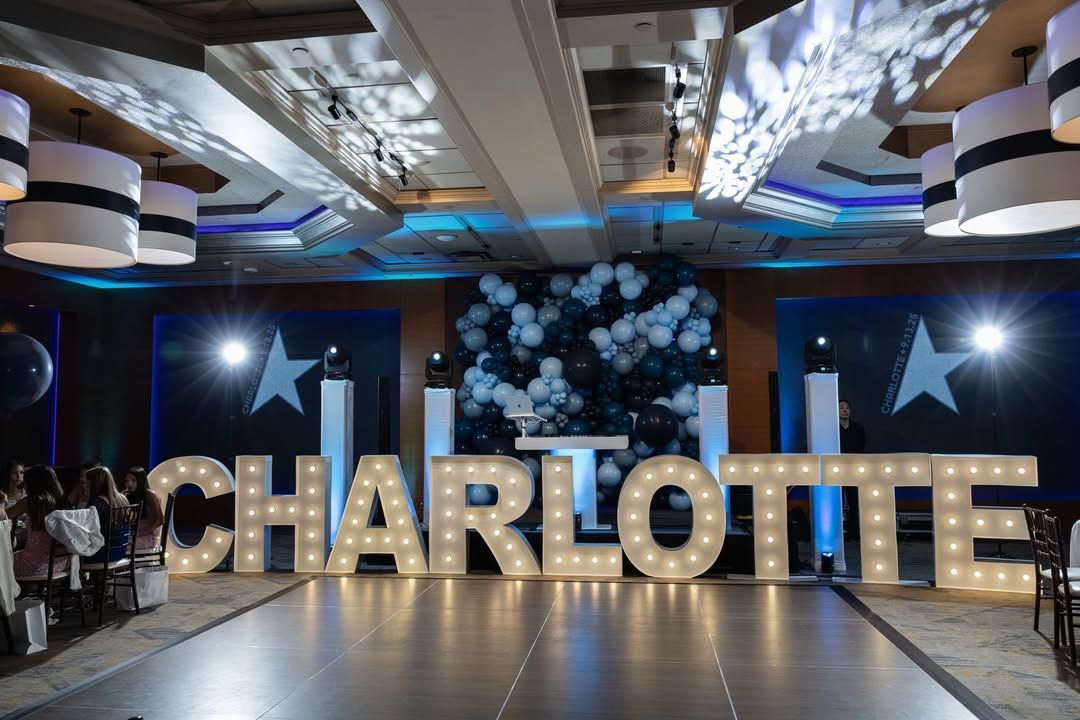 Large gold “CHARLOTTE” letters in a decorated event hall with blue lighting and white floral accents