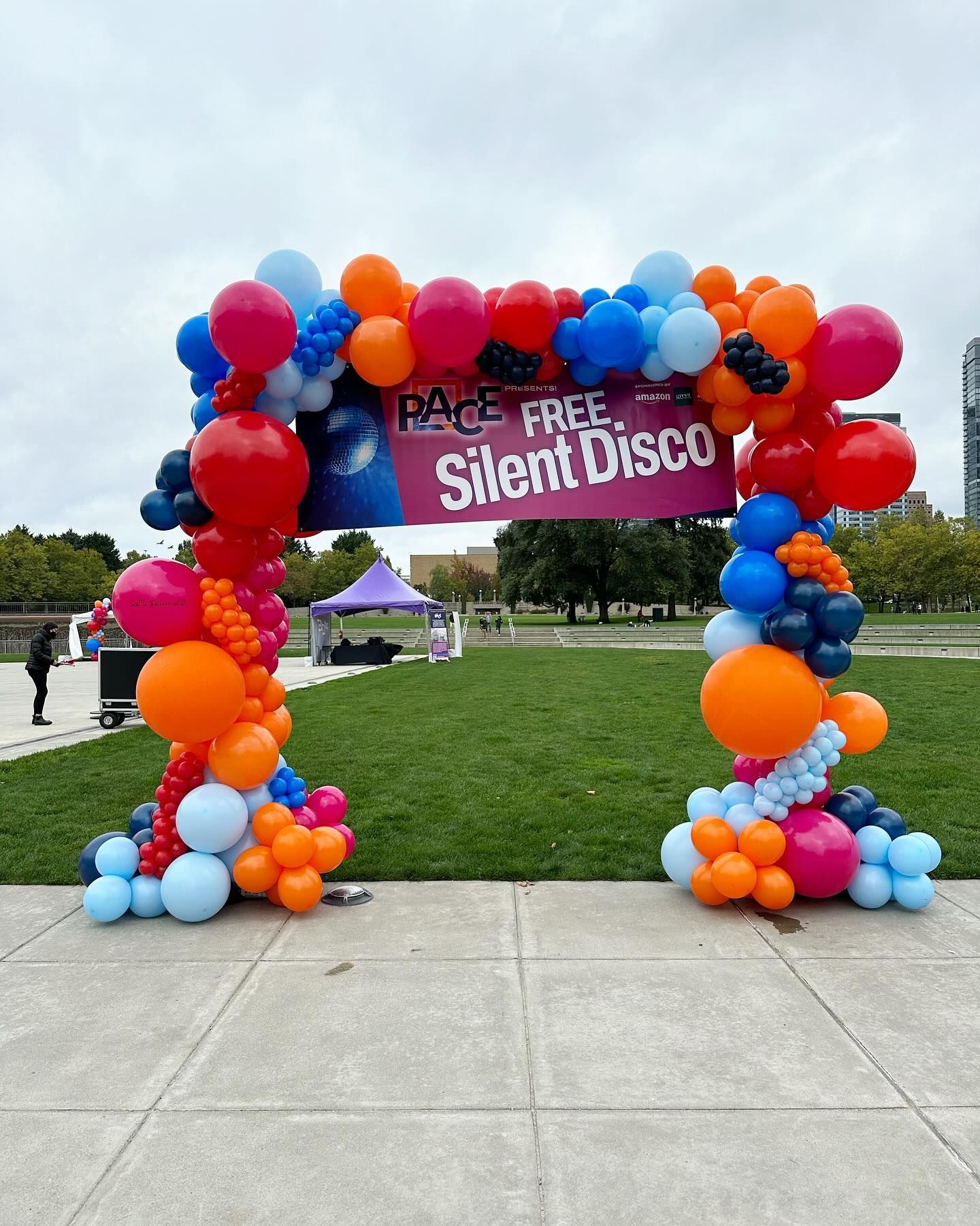 Colorful balloon arch framing a sign that reads “P.A.C.E. FREE Silent Disco” on a grassy field outdoors