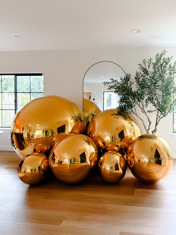 A cluster of reflective gold spheres of varying sizes arranged on a wooden floor next to a potted tree and arched mirror.