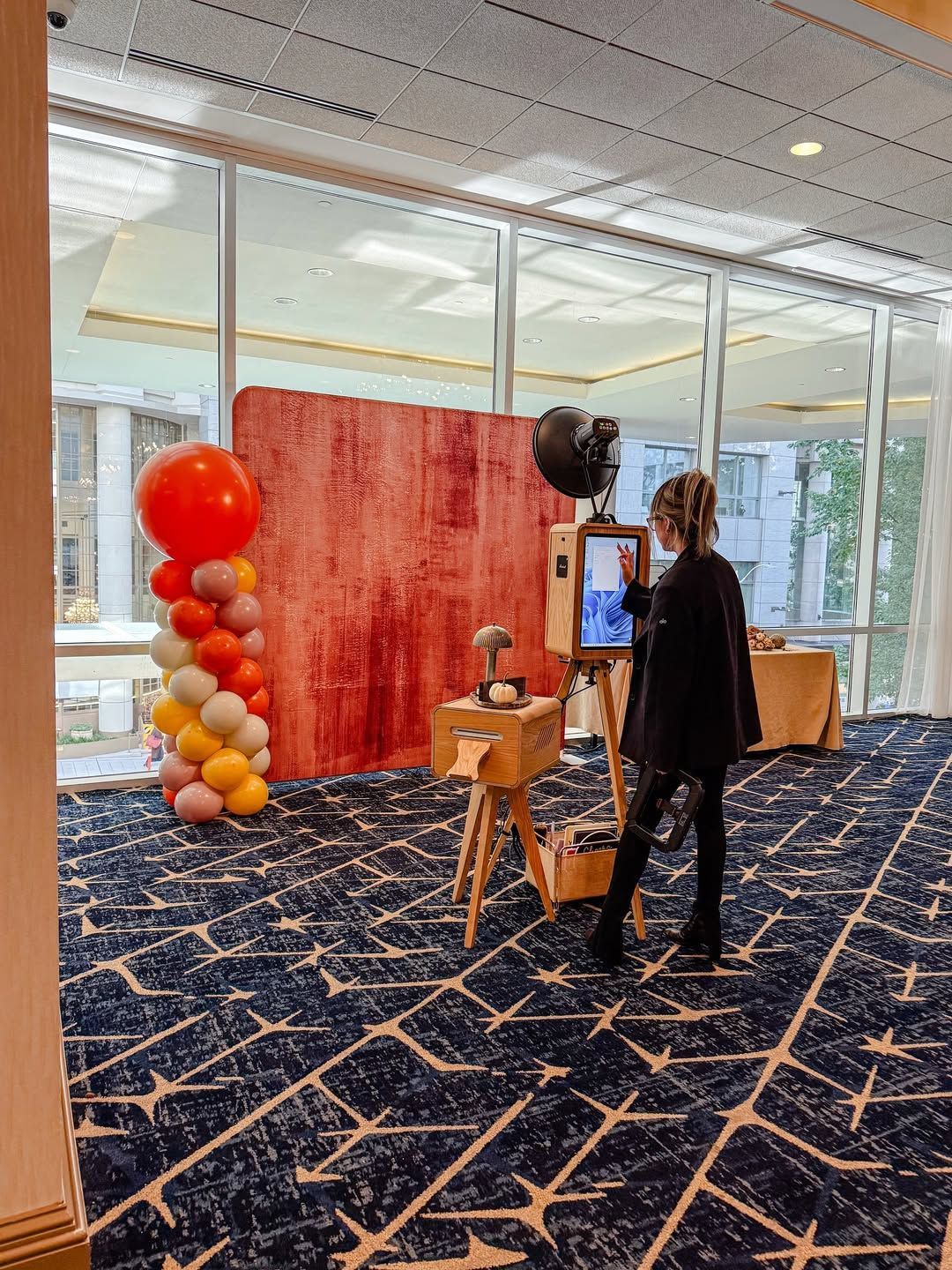 Person stands by a display booth with red backdrop, balloons, and a monitor in a bright lobby.