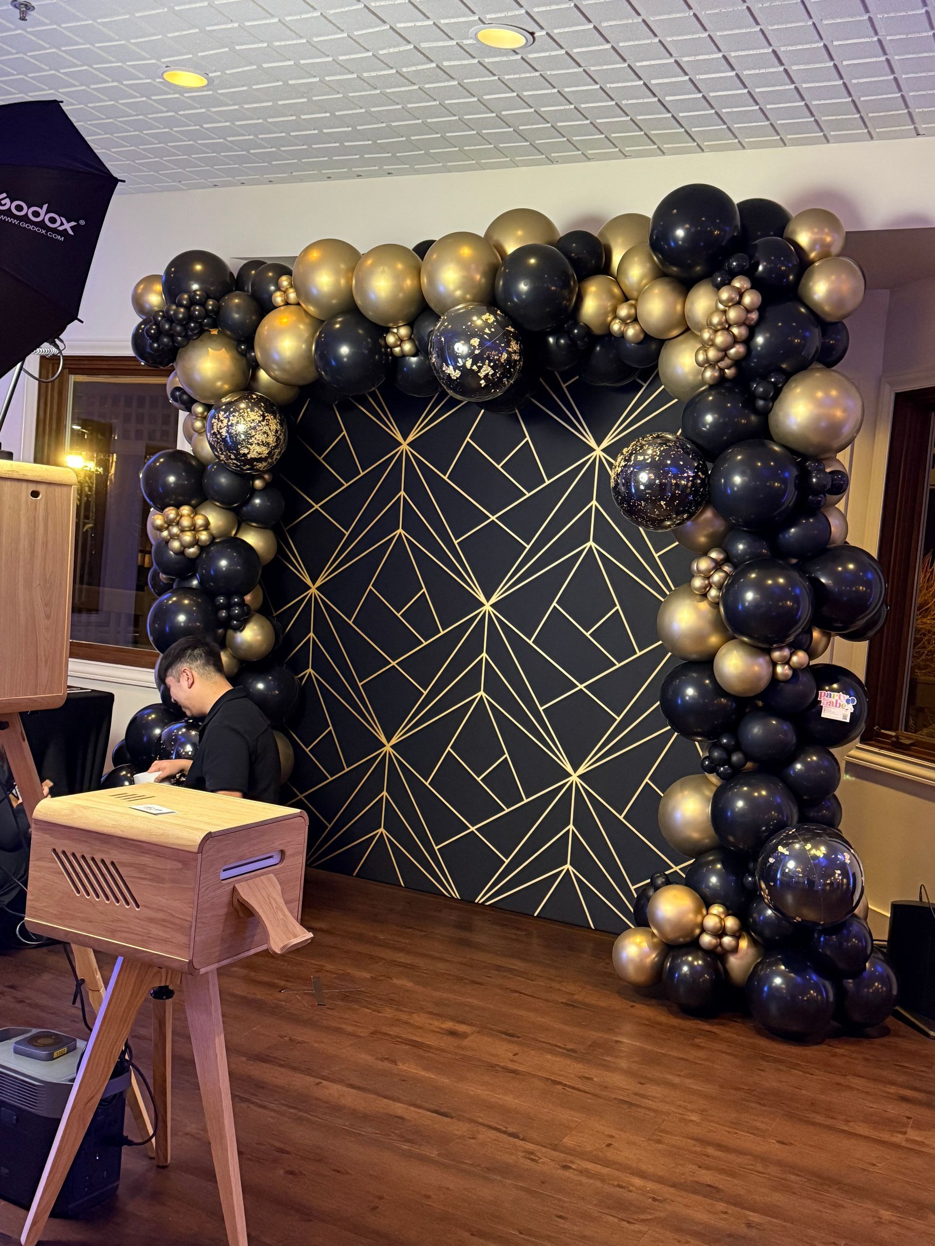 Black and gold balloon arch framing a patterned backdrop at an indoor event.