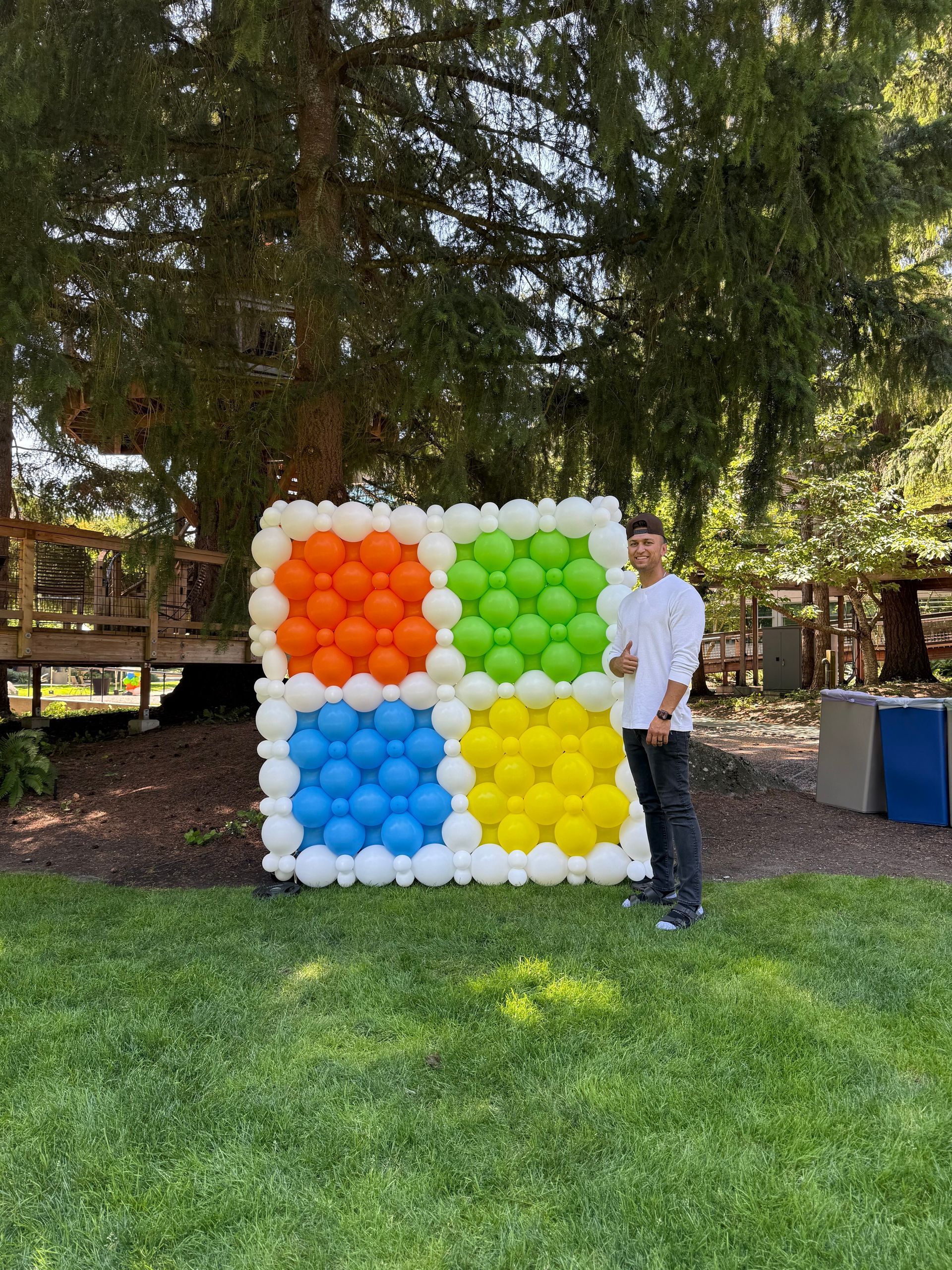Person standing beside a white balloon wall with four colored square sections outdoors under trees