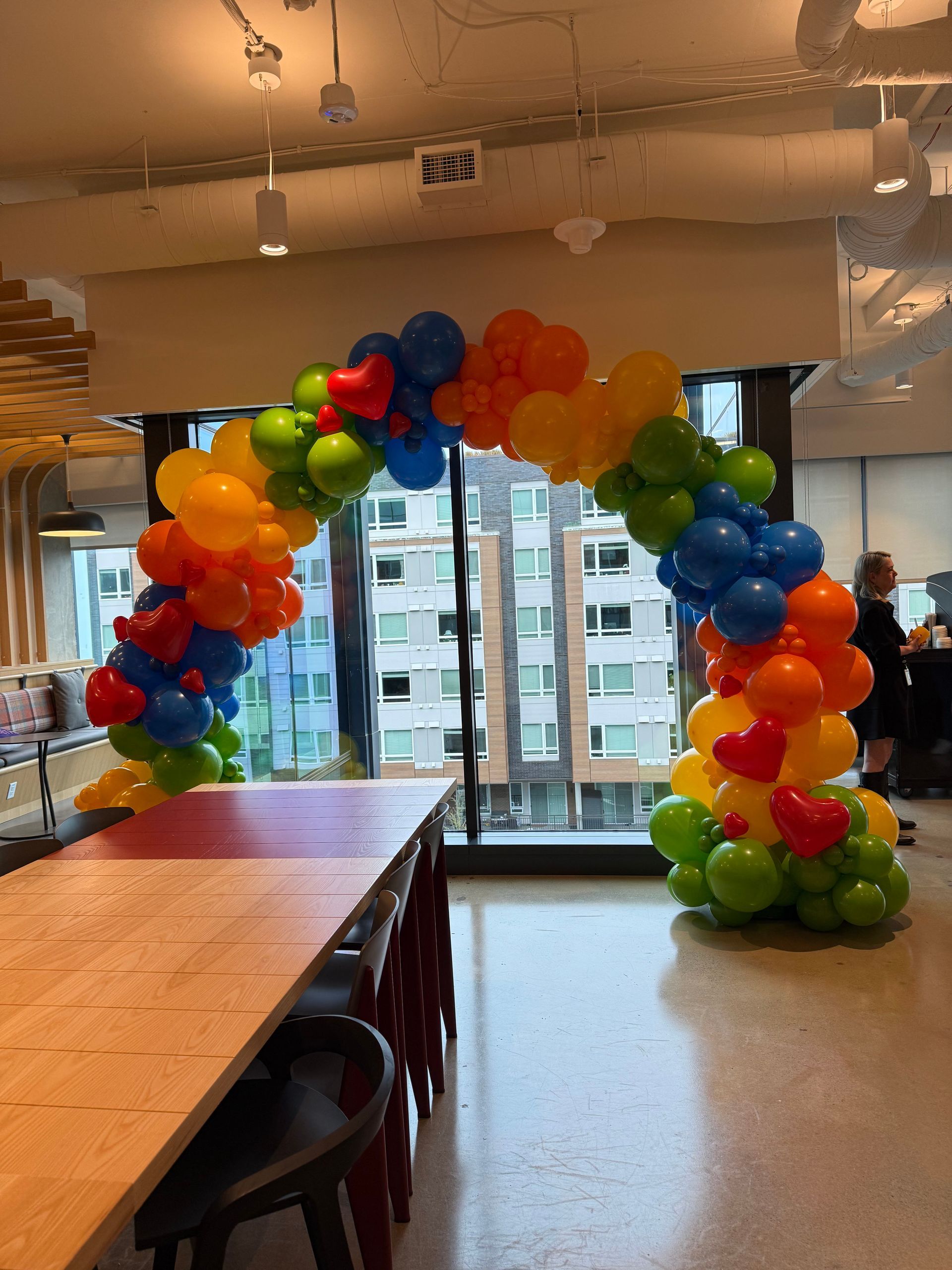 Colorful balloon arch over tables in a bright indoor room with city buildings visible through the windows.