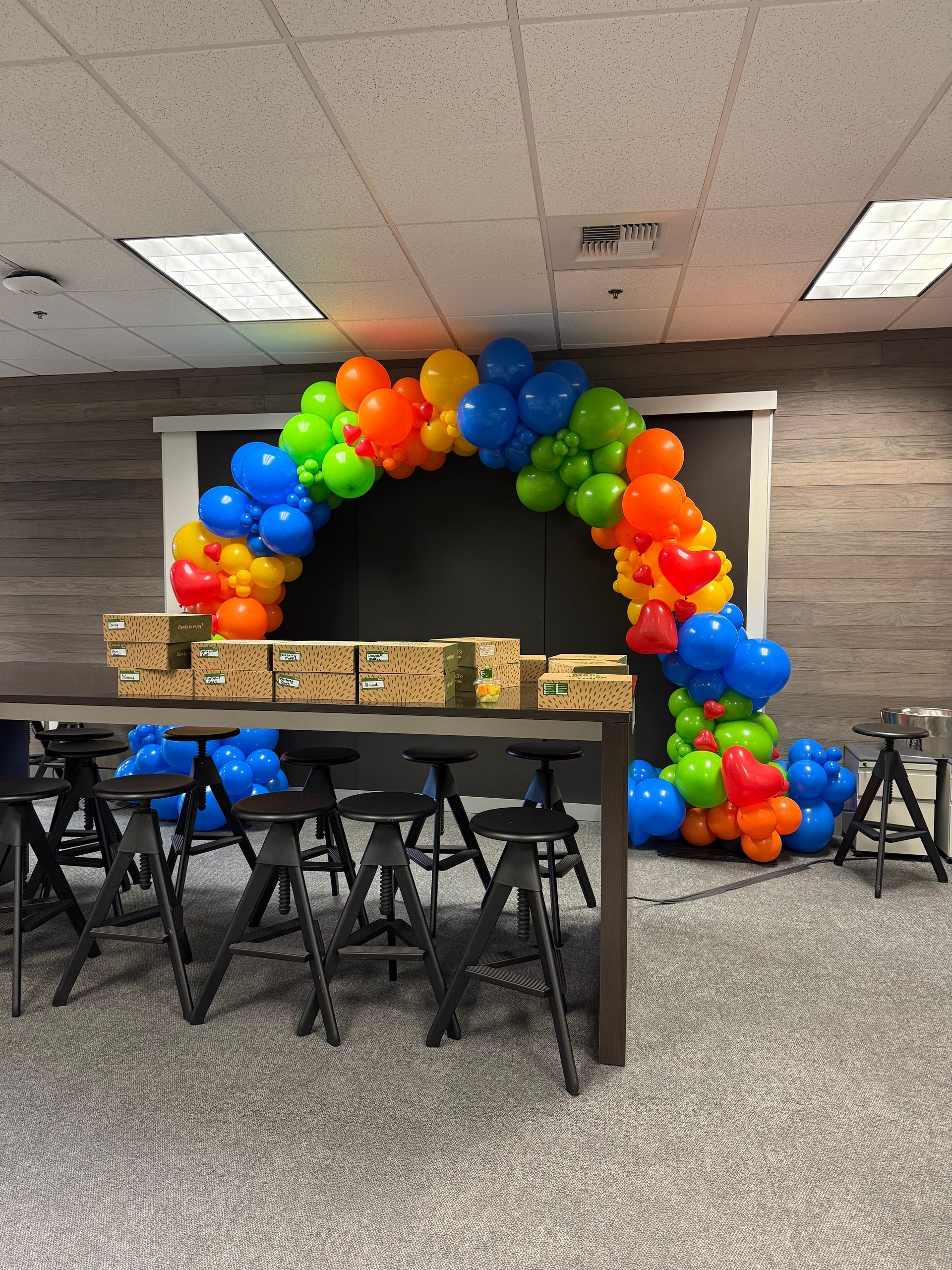 Balloon arch in rainbow colors over a table with boxes and stools in a room.