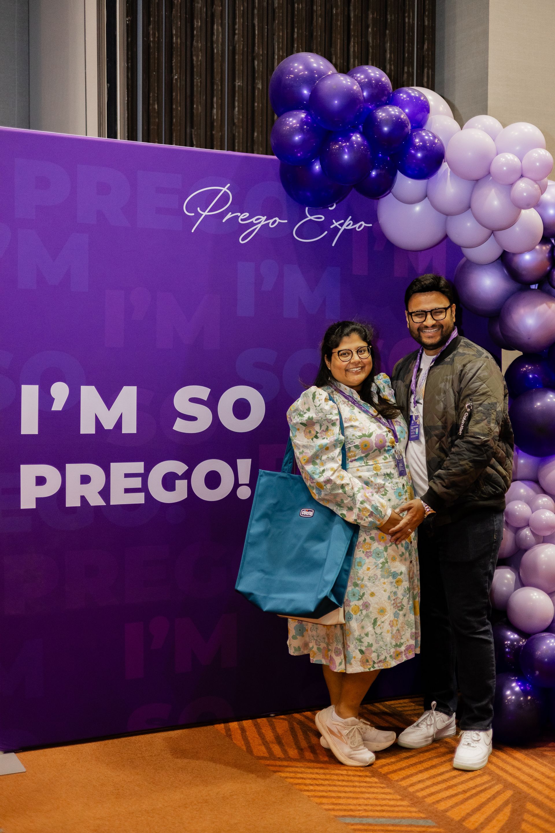 A man and a woman are standing in front of a purple wall with balloons.