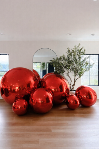 A cluster of large, shiny red spherical decorations arranged on a wooden floor next to a potted olive tree.