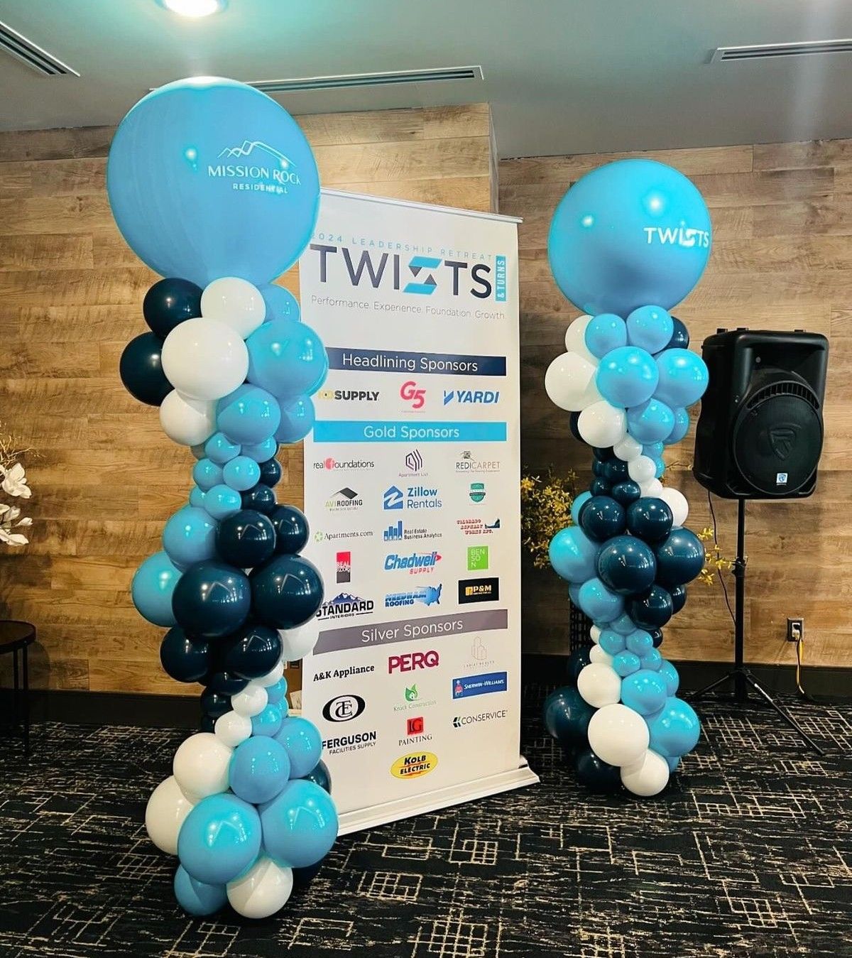 Two balloon columns with blue, white, and dark blue balloons, beside a banner that reads 