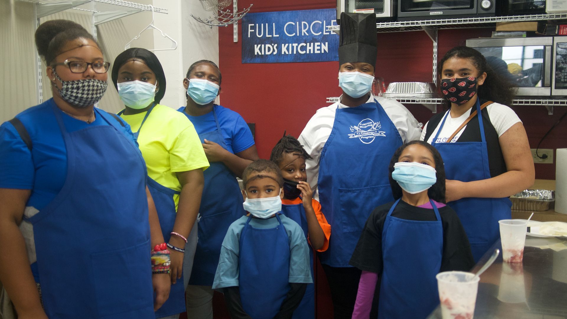 People wearing masks and aprons stand in a kitchen.