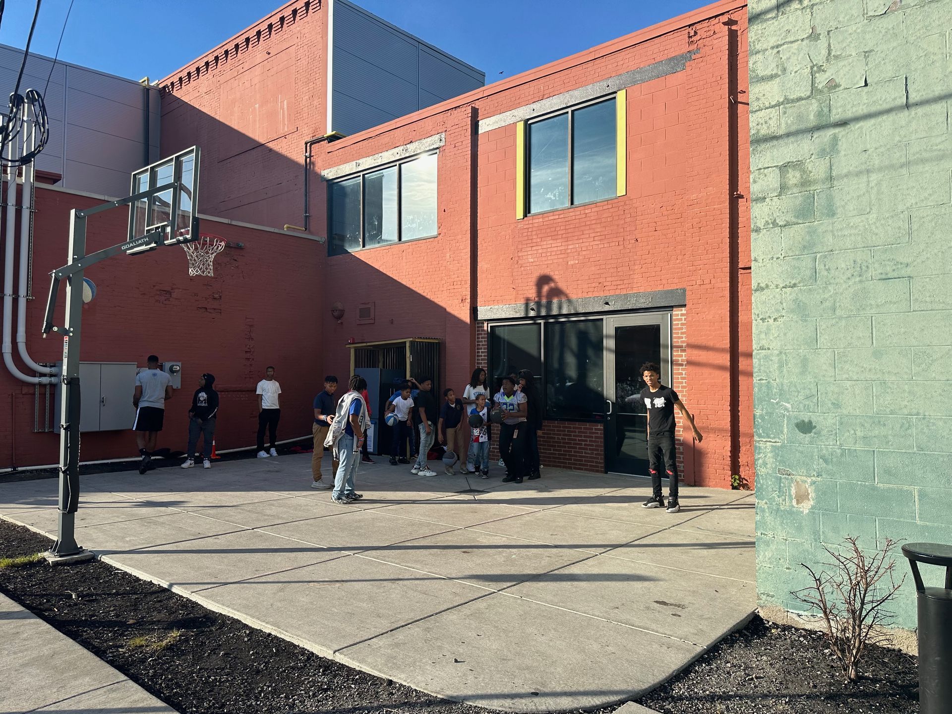 People outside a red brick building with a basketball hoop. Several are gathered by the entrance.