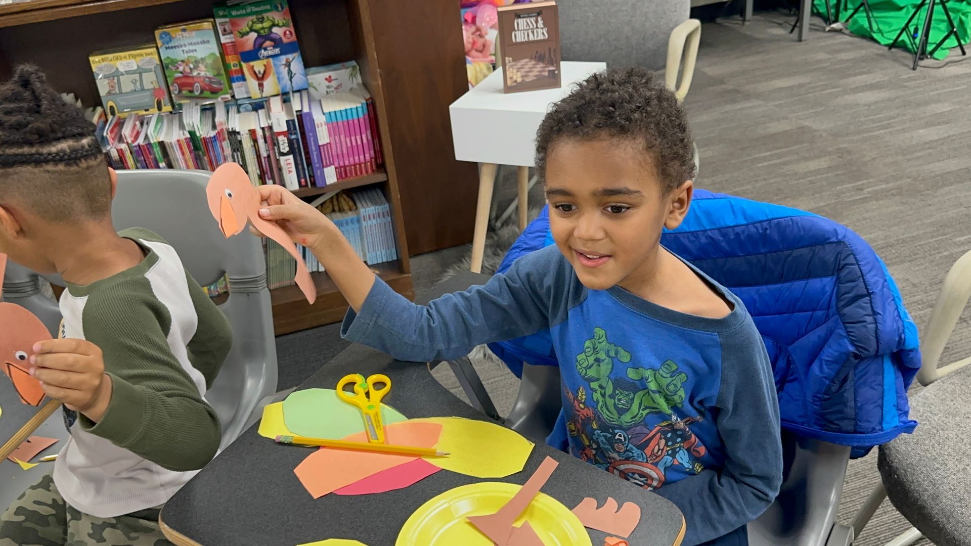 Boy at table holds up a paper craft, smiling. Others craft nearby; library setting.