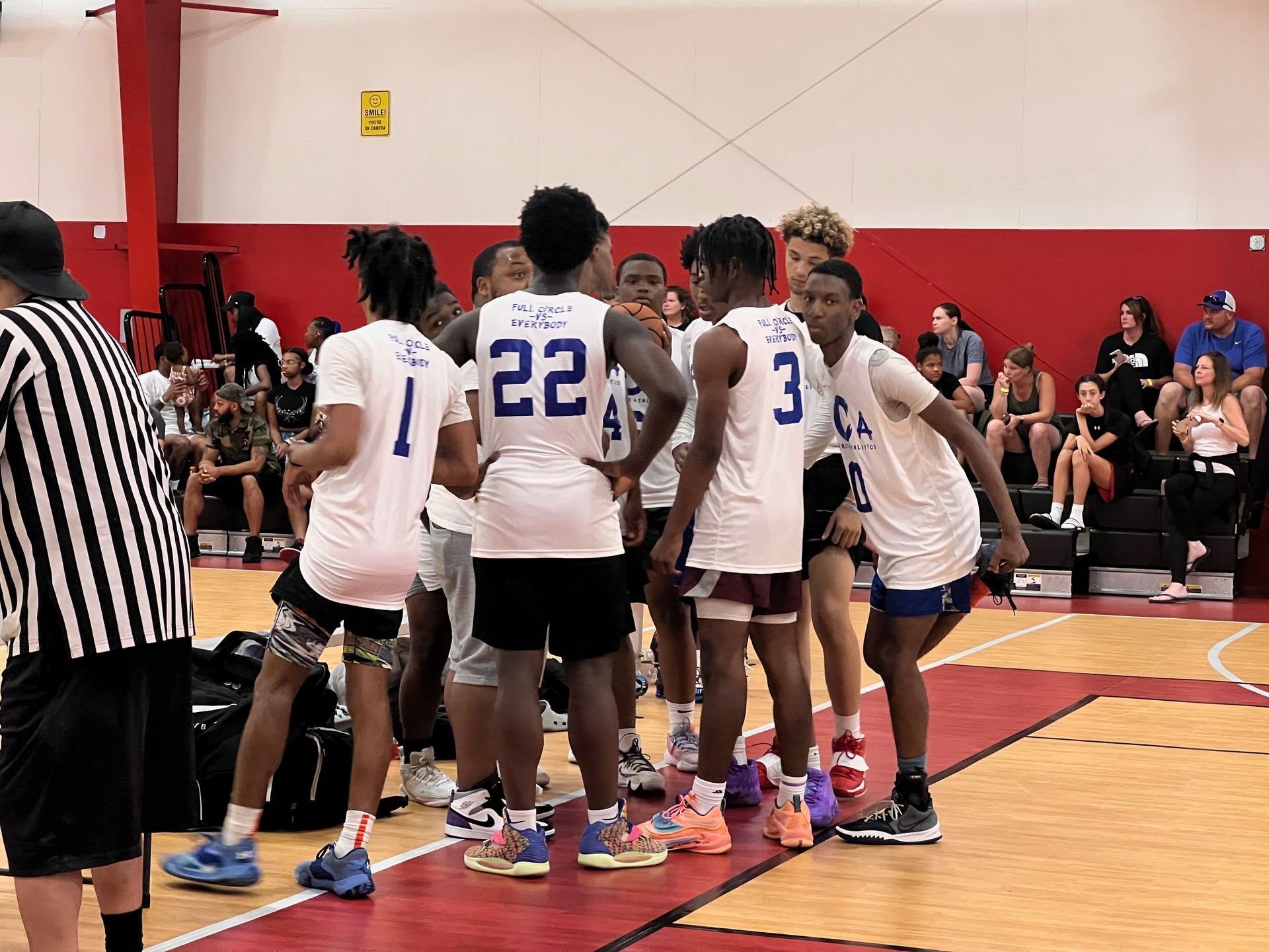 Basketball players in white jerseys huddle on a court, talking. A referee stands nearby.
