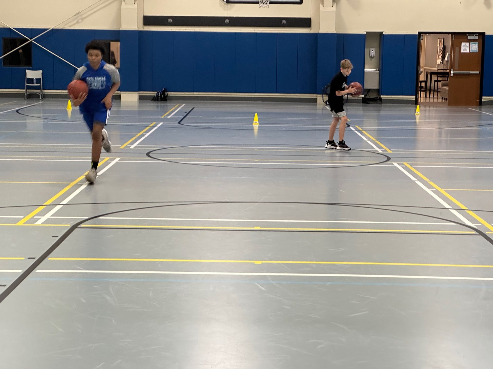 Two people dribbling basketballs in a gymnasium with blue walls and floor markings.