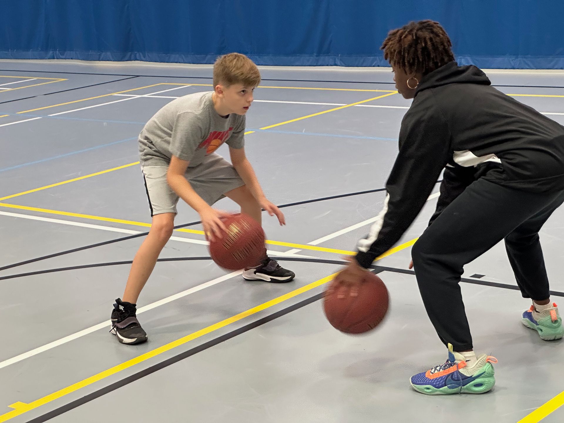 Two people dribbling basketballs on a court; one in gray and black, the other in black hoodie and pants.