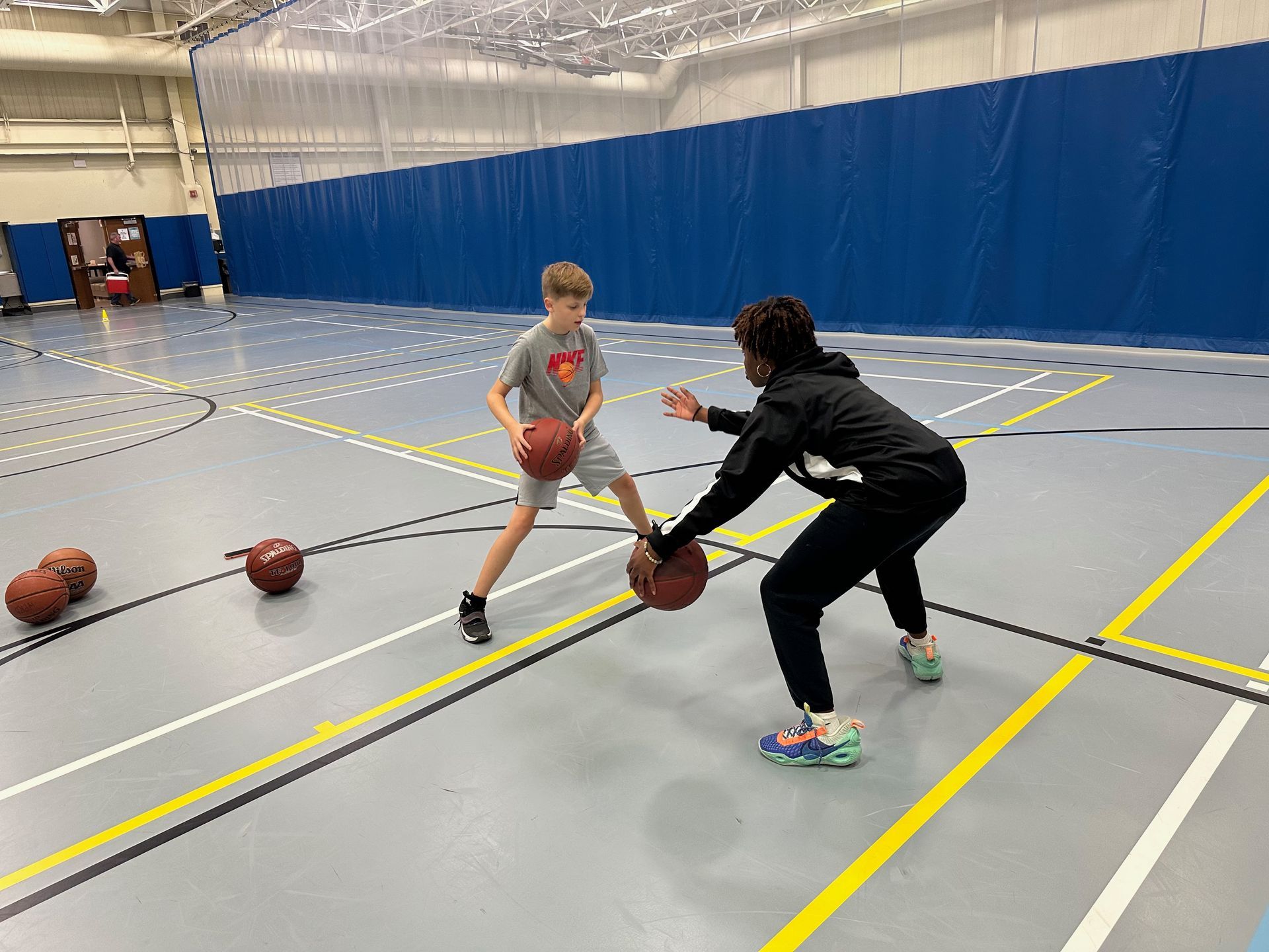 A person and a child practicing basketball dribbling in a gym with several basketballs on the floor.