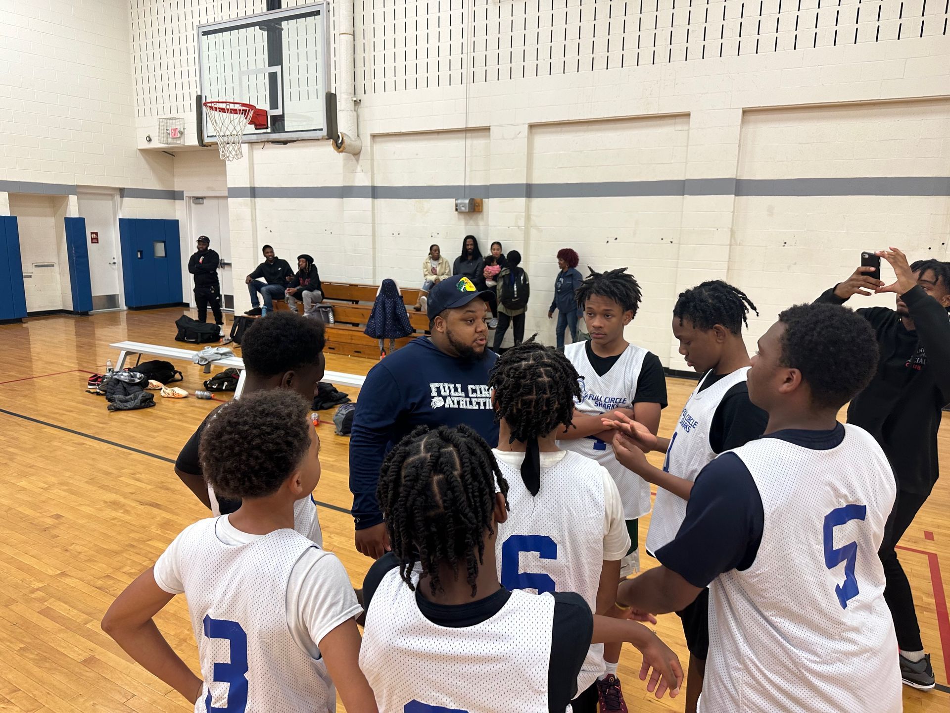 Basketball coach talking to team in gym. Players wearing white and blue jerseys, huddled together.