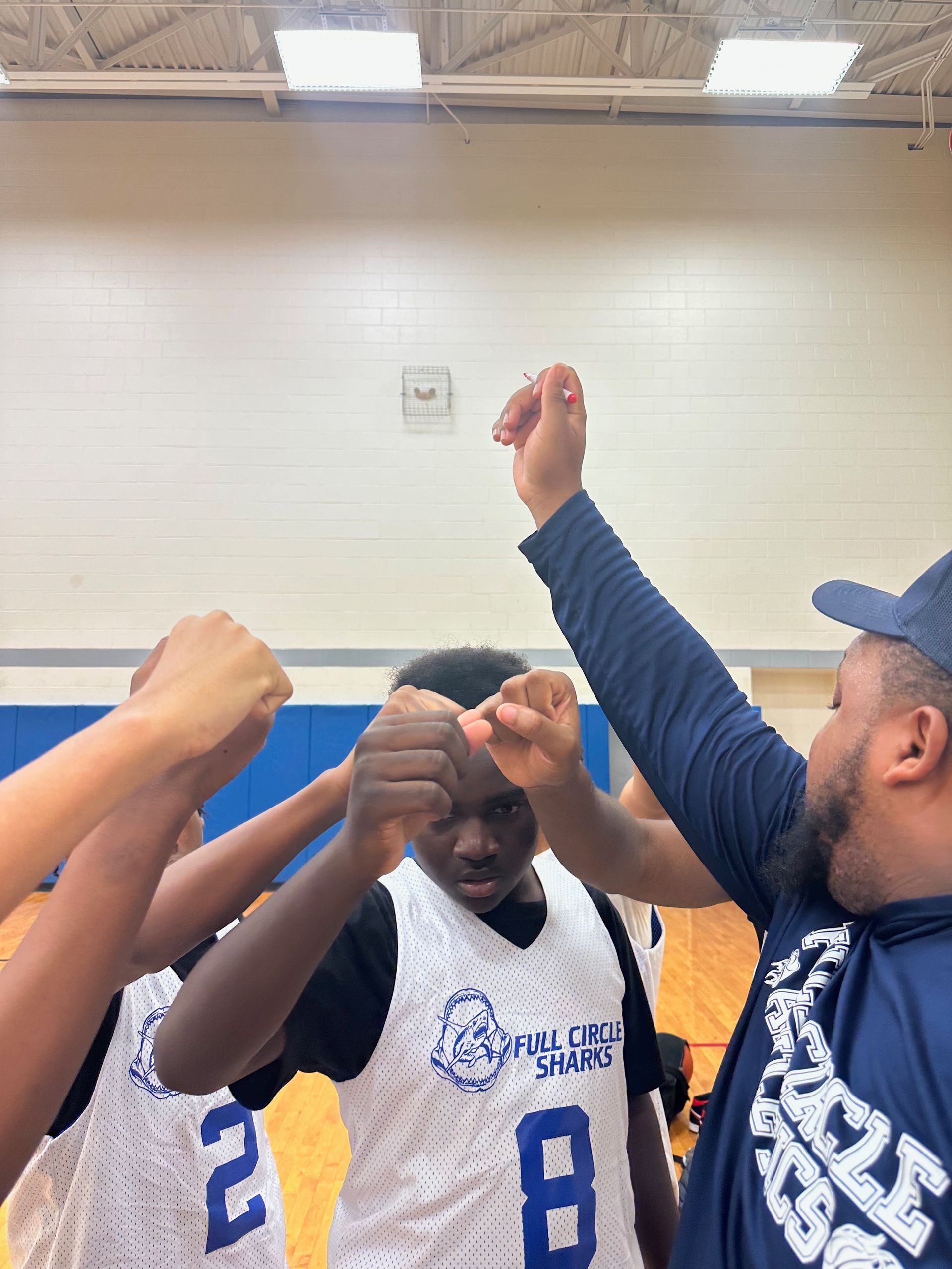 Basketball team huddles, arms raised in unity. Players wearing blue and white jerseys, coach with beard.
