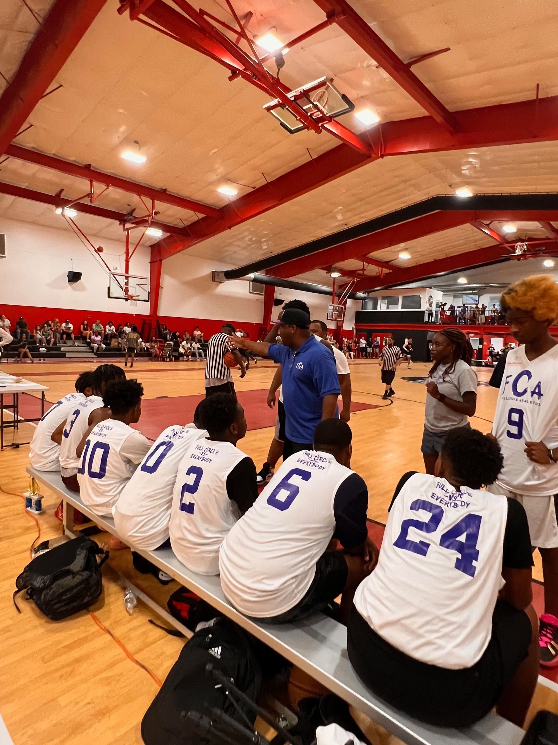 Basketball team on bench listening to coach during a game. Gym setting, players wearing white jerseys with blue numbers.