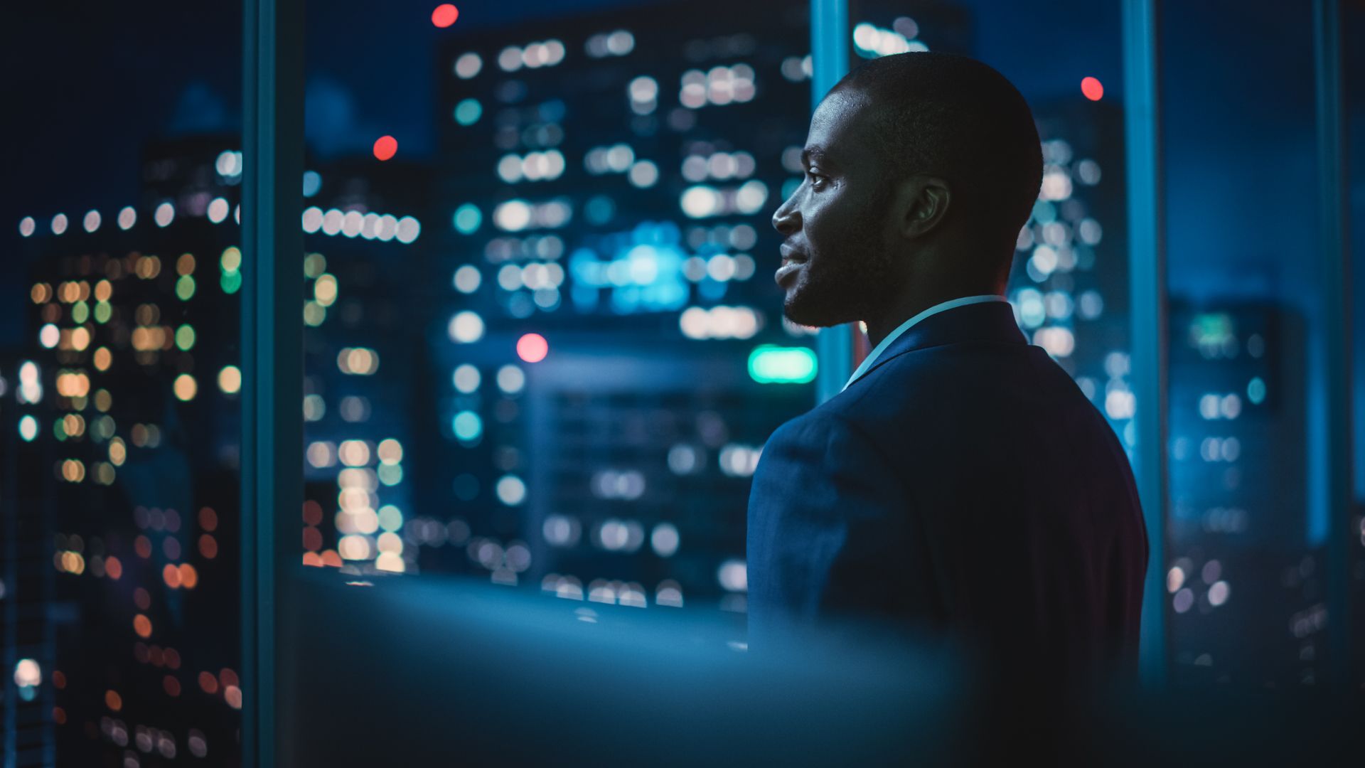 Man in suit looks out at city lights from a high window at night.