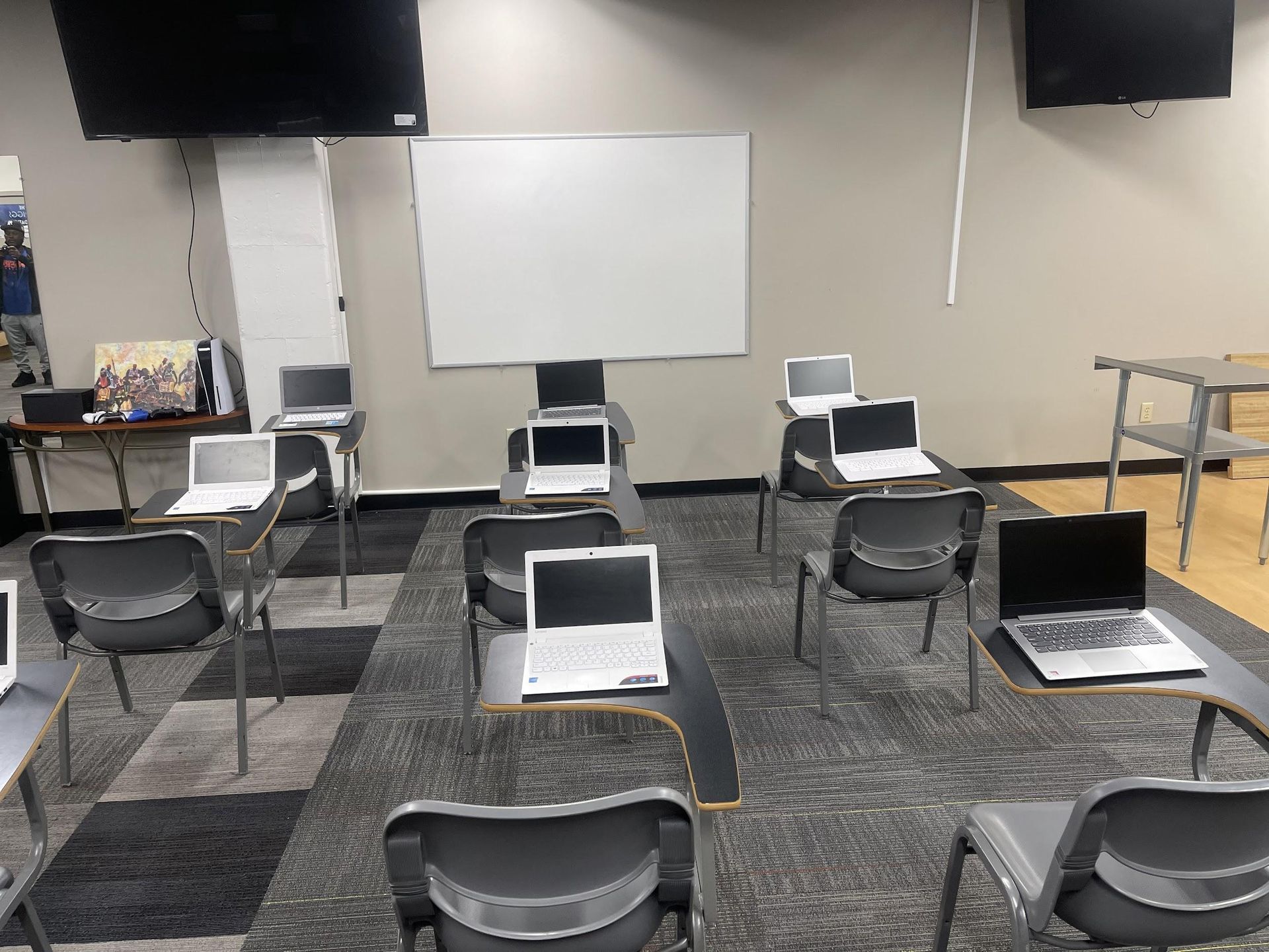 Classroom with laptops on desks, arranged towards a whiteboard and two TVs. Gray chairs and carpet.