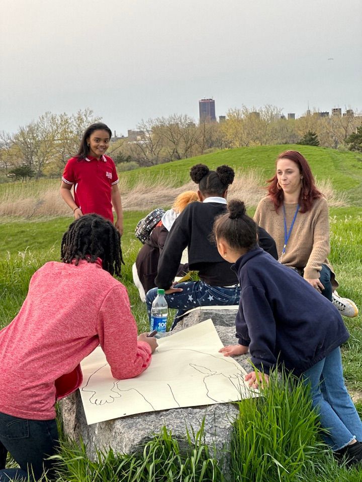 Group of people drawing on a large sheet of paper outdoors. A woman observes.