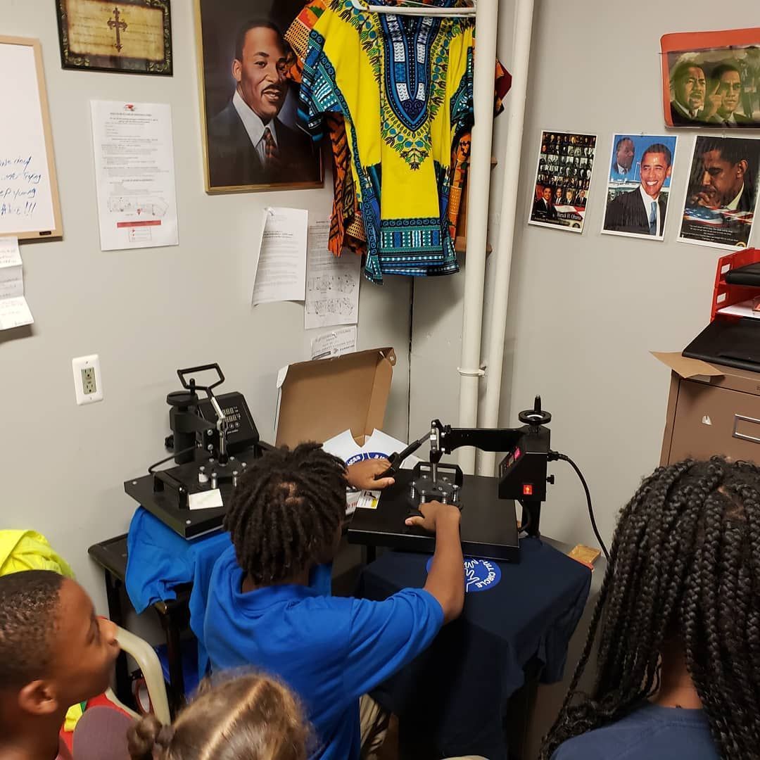 Children watch a person at a heat press machine in a room with a portrait of Martin Luther King Jr. and other Black figures.