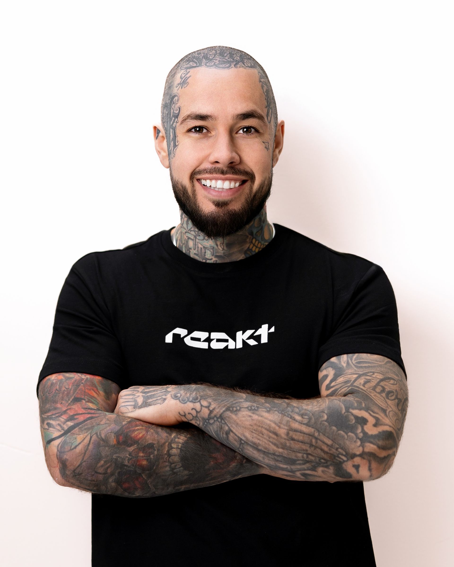 A smiling, heavily tattooed person with a shaved head and beard, wearing a black t-shirt with arms crossed against a wall.