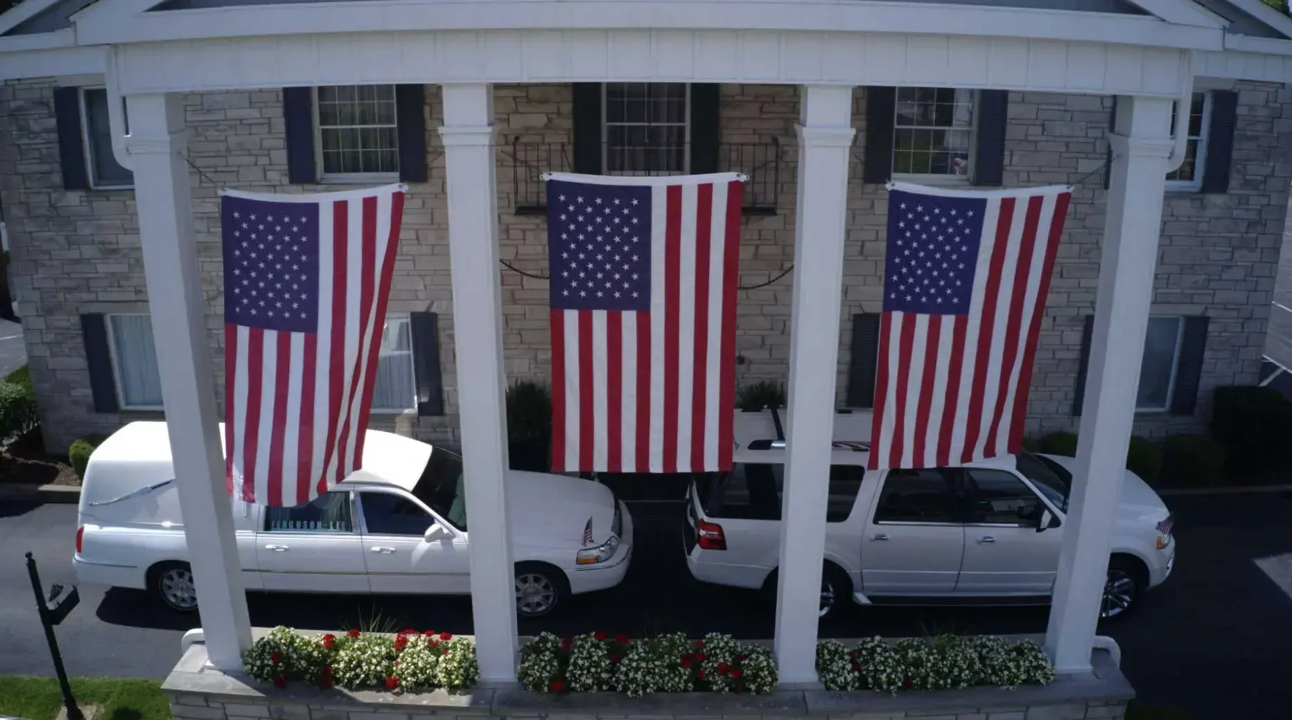 Three large American flags hang from a porch over parked white vehicles.