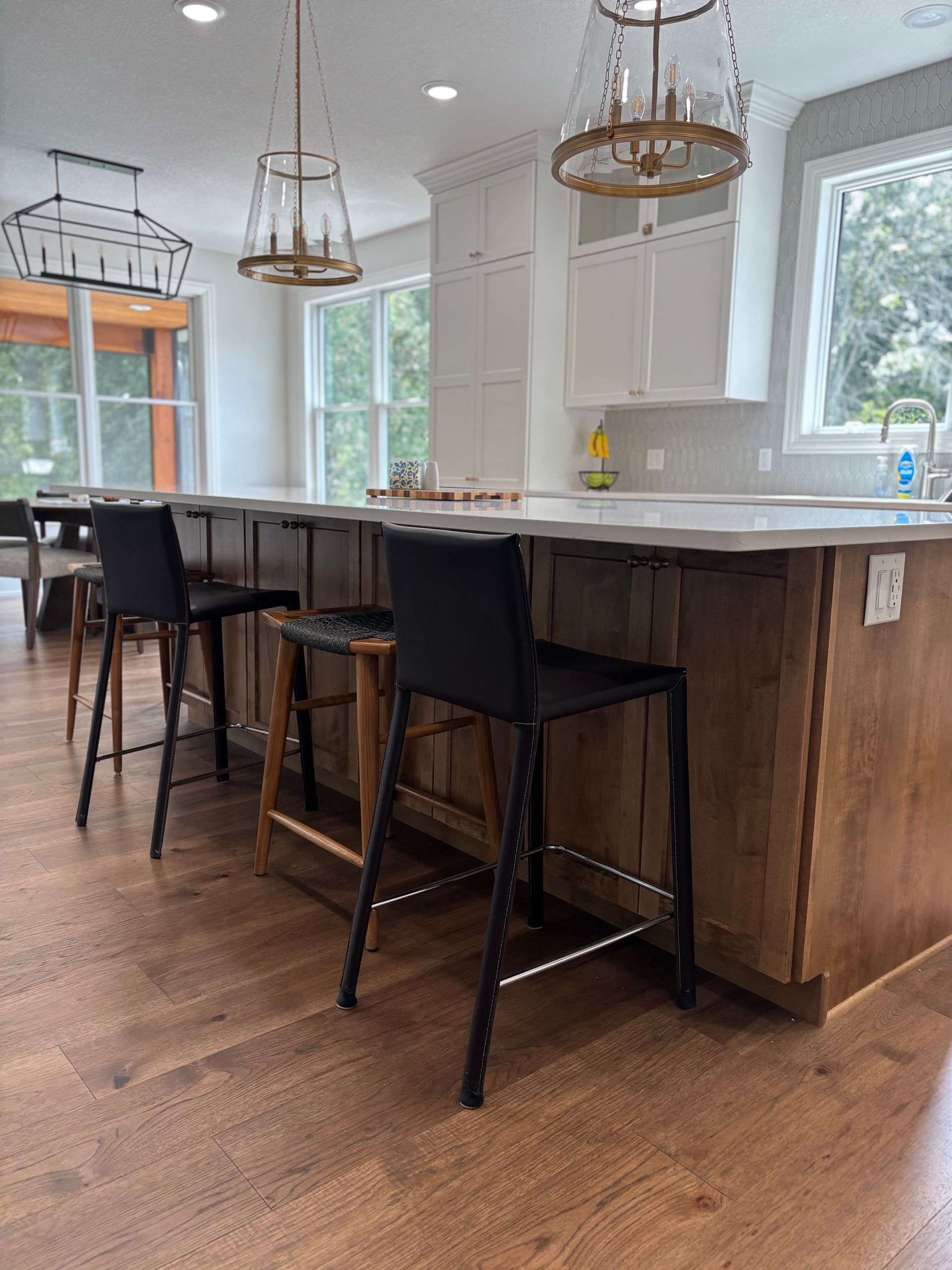 Modern kitchen with island, brown cabinets, white countertop, black bar stools, wood floor.