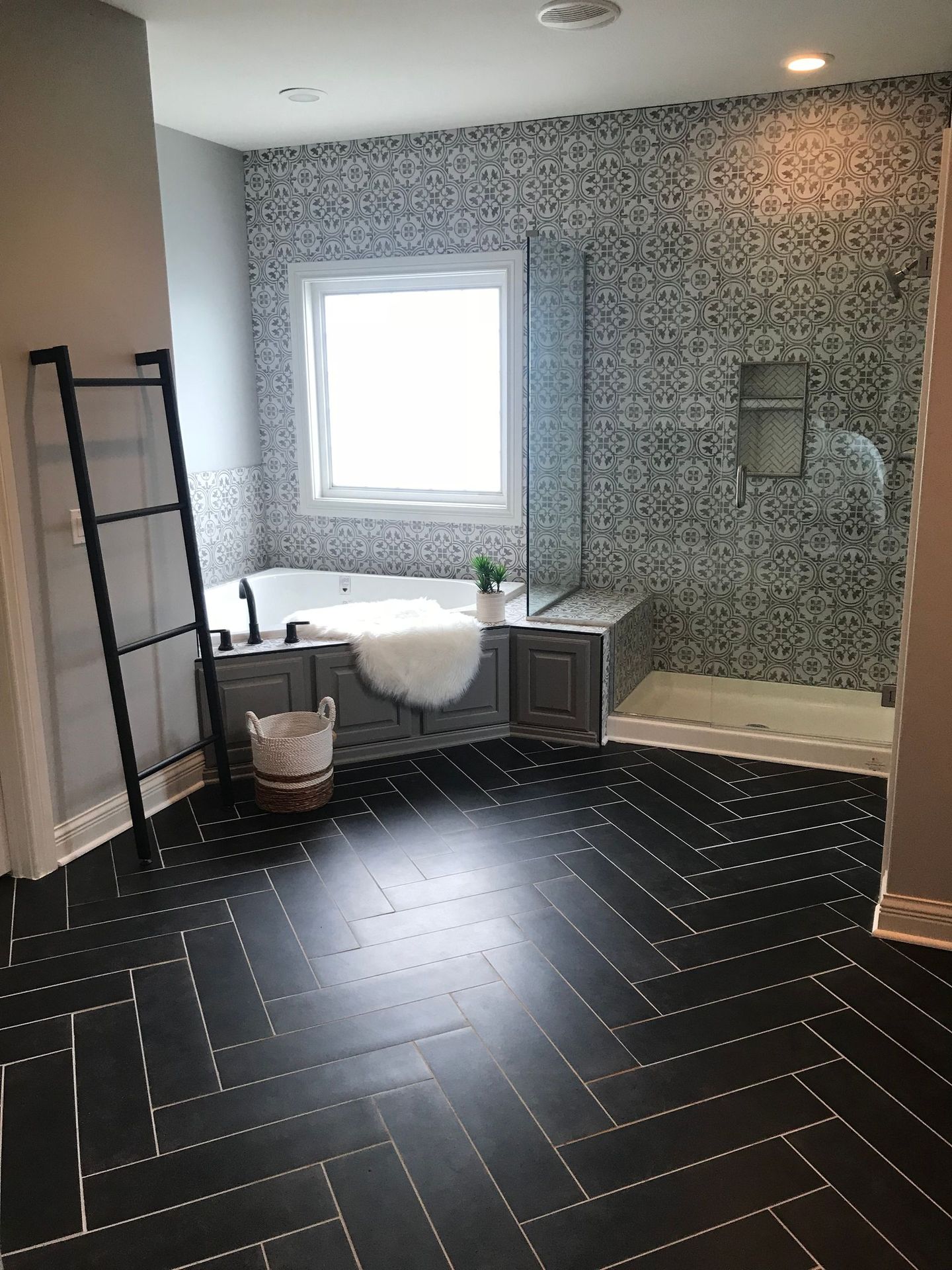 Bathroom with black herringbone floor, gray cabinets, mosaic tile wall, window, tub, shower.