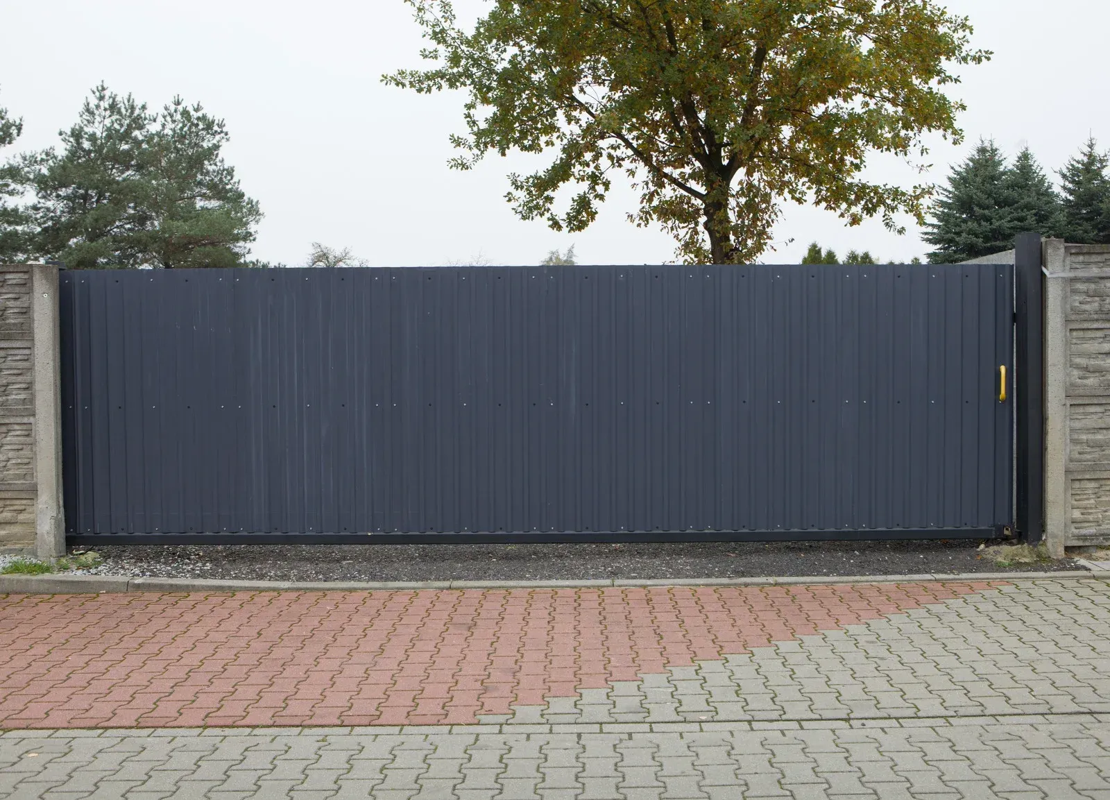 A gray sliding gate is sitting on a brick driveway next to a tree.