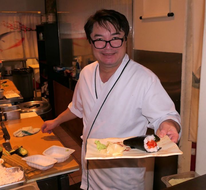 Chef in a kitchen holding a plate with sushi and vegetables, wearing a white coat and glasses.