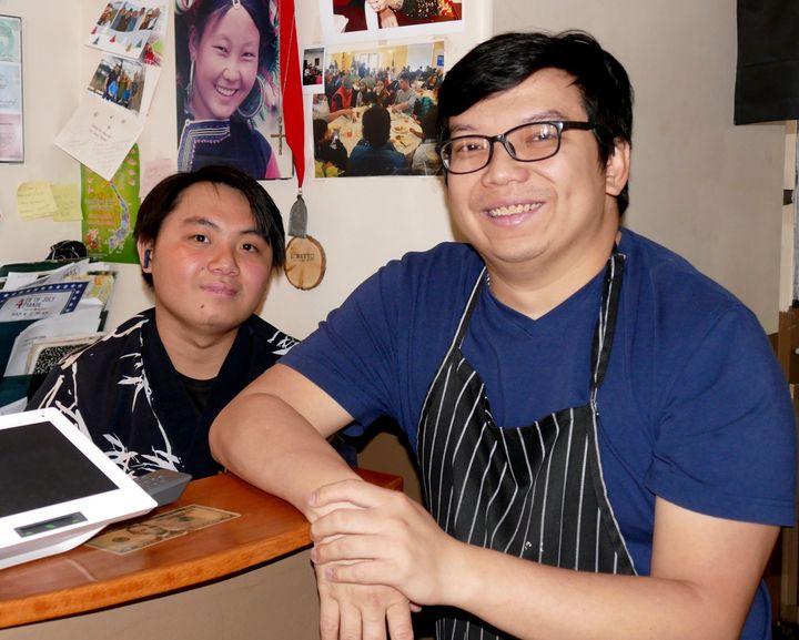 Two people smiling at a table in a casual indoor workspace, with photos and papers on the wall behind them.