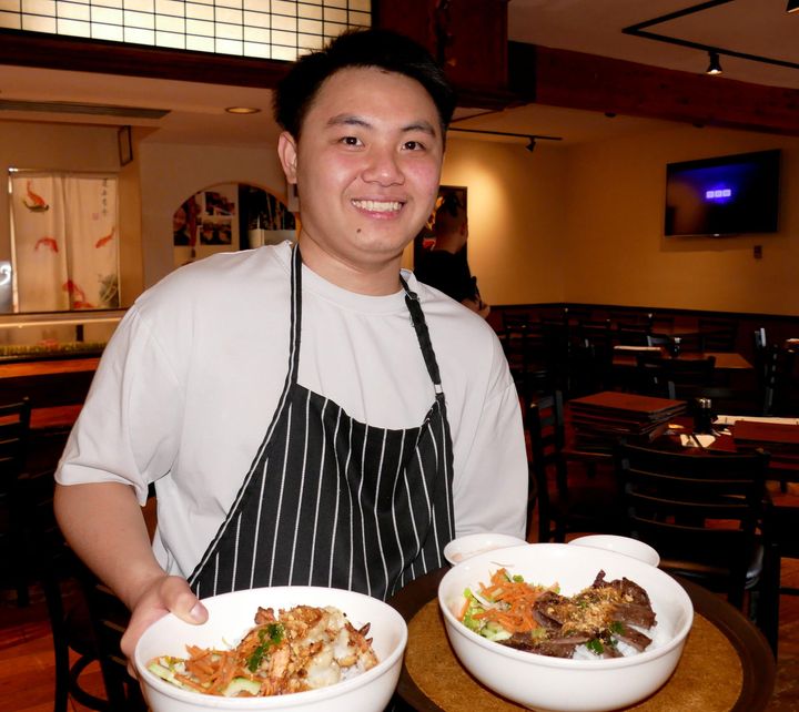 Chef in a striped apron smiles while holding two bowls of food in a restaurant kitchen