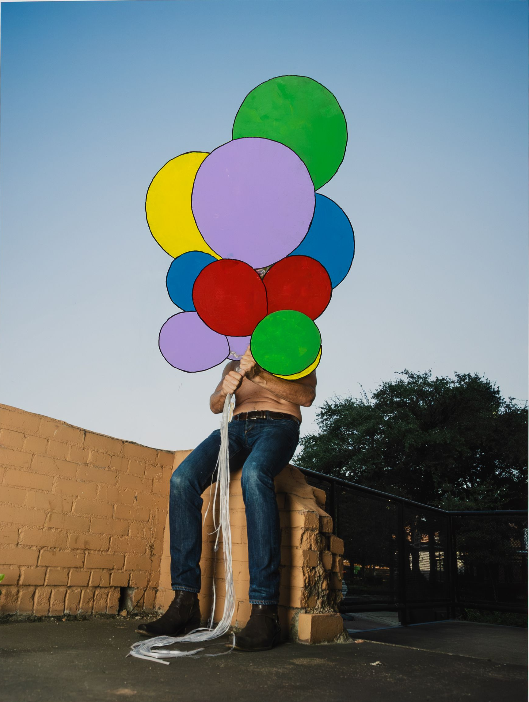 shirtless guy sitting on brick holding a bouquet of balloons in front of his face