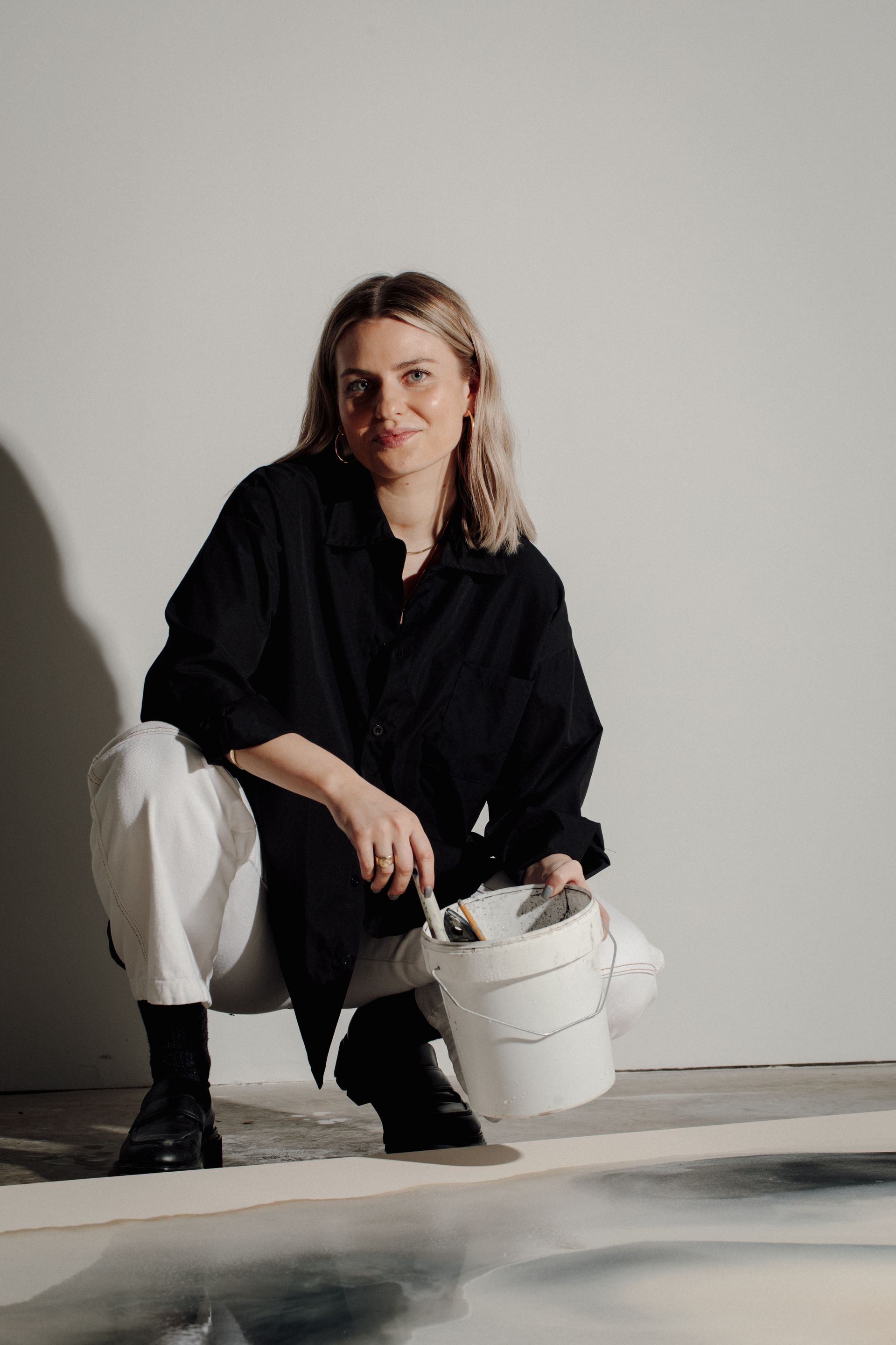 Joy Kinna crouching over a painting as she works on it. She looks into the camera and smiles with paintbrush and water bucket in hand. shot by Brit Gill Photography