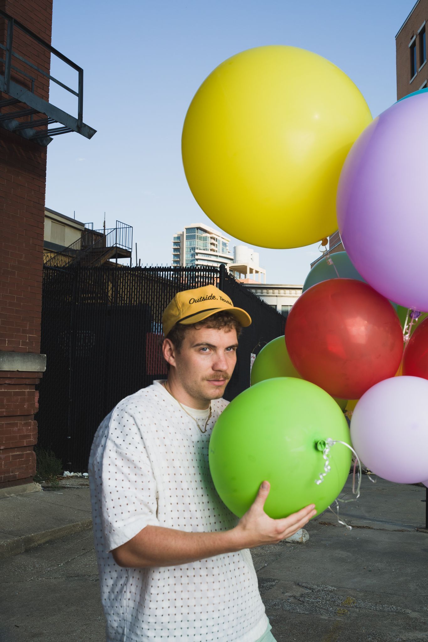 Nicholas Sanders (artist) holding a bouquet of colorful balloons posing for portrait photo, scenic background of a city