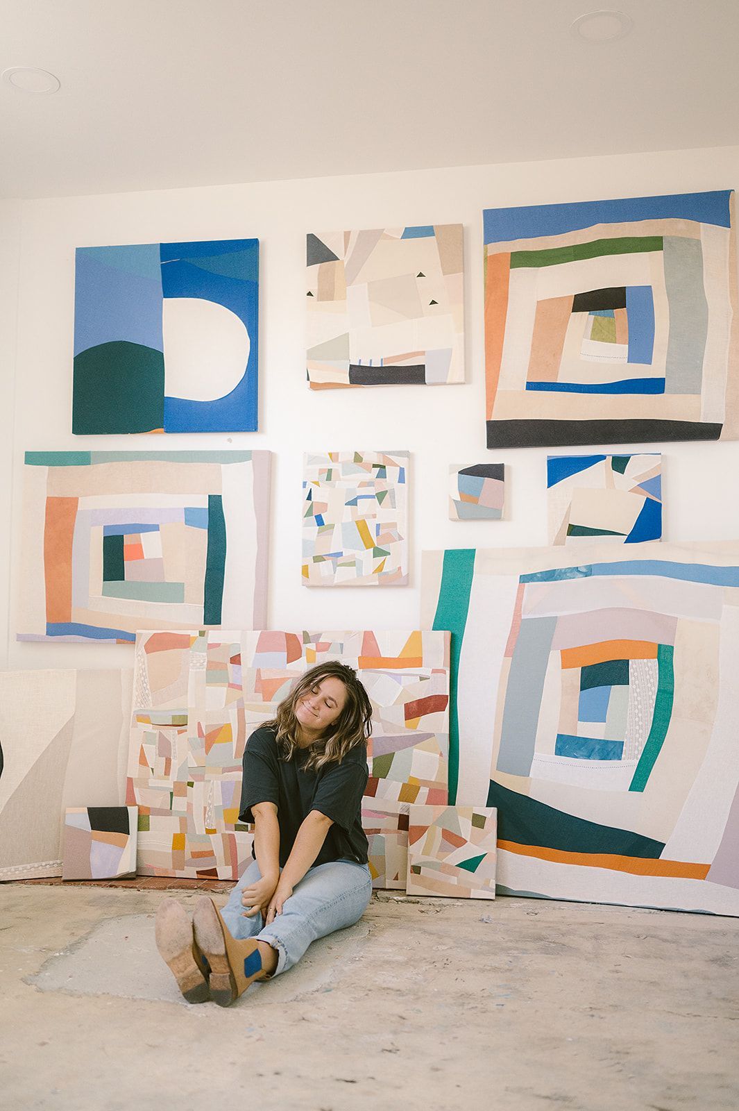 Sydney in her studio with her art hanging on the wall behind her. she sits on the floor and tilts her head to the side and smiles softly.