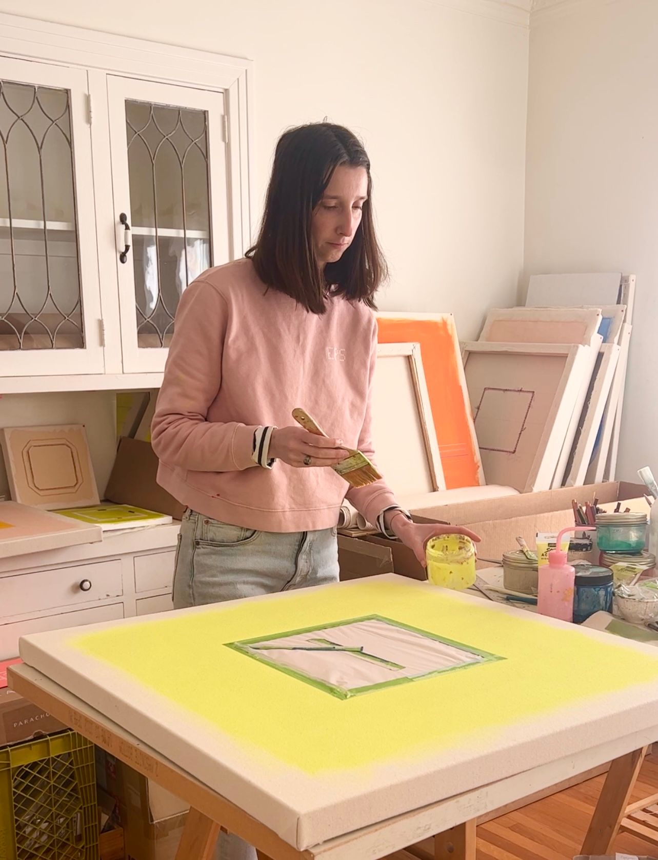 Emily Keating Snyder in her studio painting a piece with a vibrant yellow/green. The paintbrush is in one hand and her paint can is in the other. She is wearing a pink sweatshirt with her initials on it and finished works behind her.