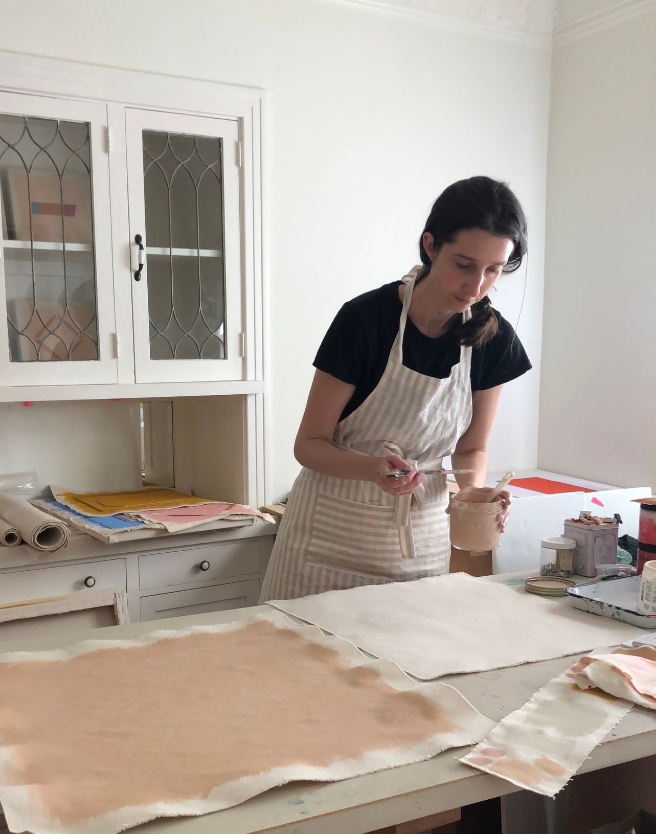 Emily Keating Snyder in her studio painting loose canvas with a peachy pink color. She is wearing an apron and leans over the canvas with brush in hand
