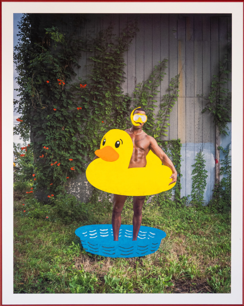 Undressed male with a yellow duck floatie worn around his waist with yellow goggles standing in a blue small pool, located in a backyard