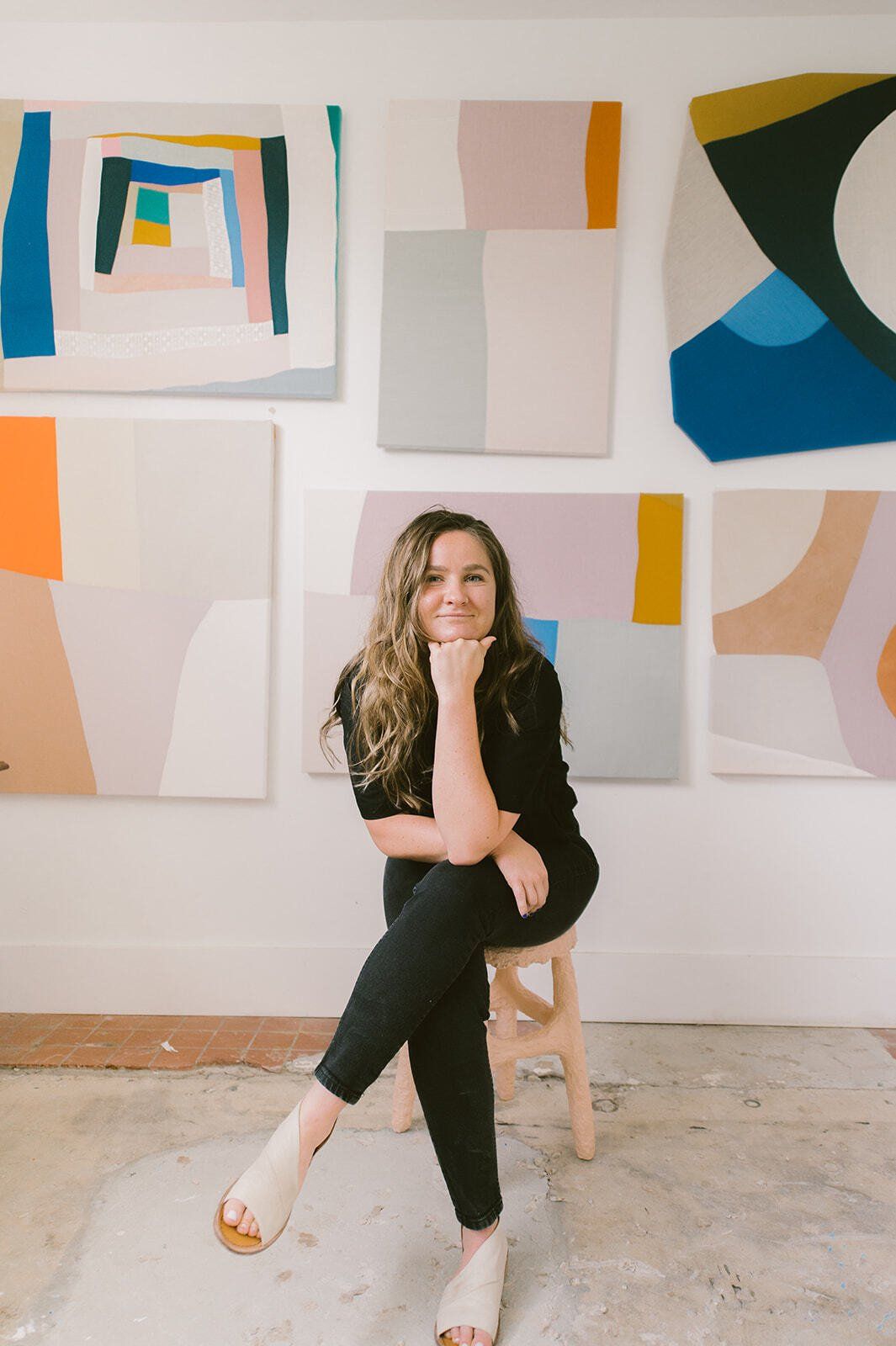 Sydney in her studio with art behind her. she is sitting on a stool, resting her elbow on her crossed legs and propping her head up, smiling softly.